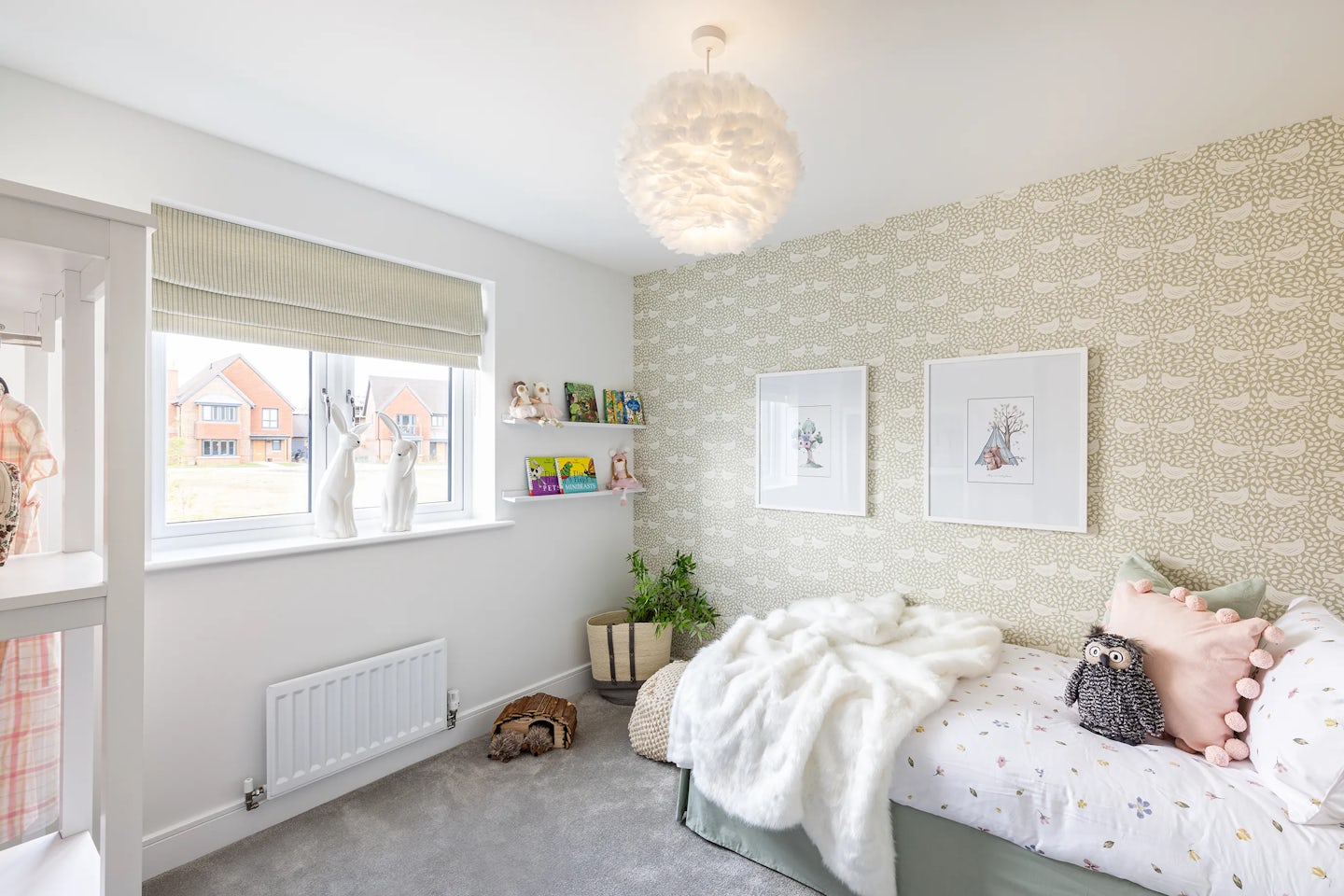 Cozy, light-filled bedroom featuring a single bed dressed in a white fluffy blanket and assorted pillows, including a playful owl design. A cloud-like pendant light hangs above, enhancing the whimsical atmosphere. The wall behind the bed is adorned with subtle patterned wallpaper and two framed prints. Beneath the beige window blind, soft toys and books rest on the windowsill, adding charm and personality. A white radiator sits below the window, and the grey carpeted floor hosts a woven basket and small plant
