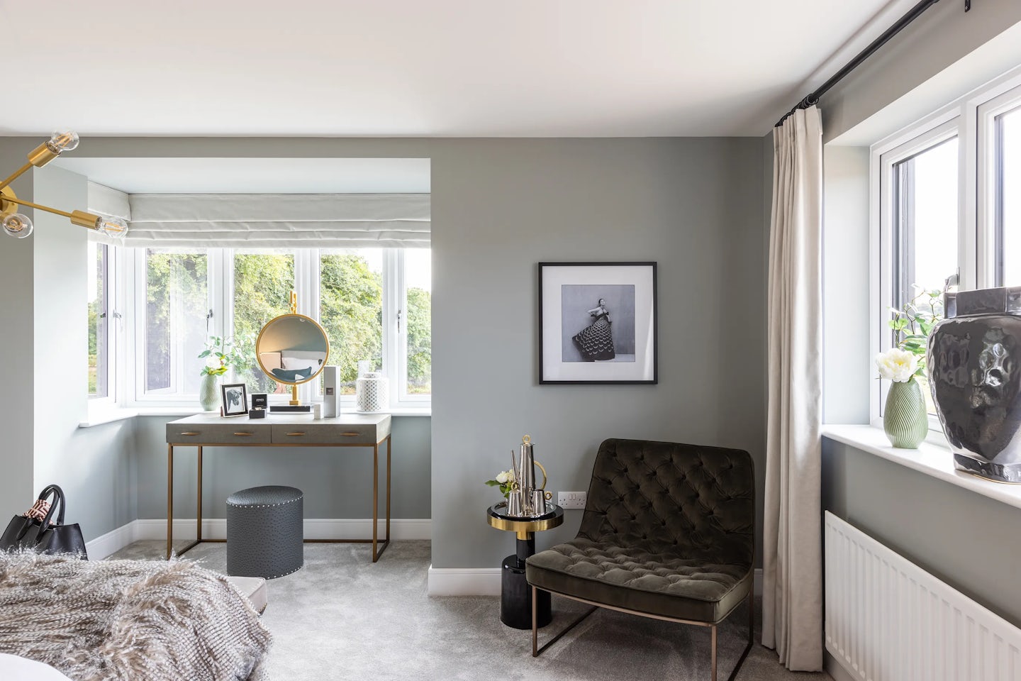 Elegant bedroom workspace featuring a pale wooden desk with slim gold legs and drawers, positioned in front of a wide window with white blinds. The desk is adorned with a round tabletop mirror, framed photos, decorative jars, and a potted plant, suggesting personal touches and daily ritual. To the right, a green velvet armchair and small round table with a brass lamp and greenery create a cozy reading nook beneath a black-and-white wall photograph. A second window with sheer white curtains frames the far right corner, with a large black vase and white floral accent on the sill. The grey walls and carpet contribute to the soft, neutral ambiance