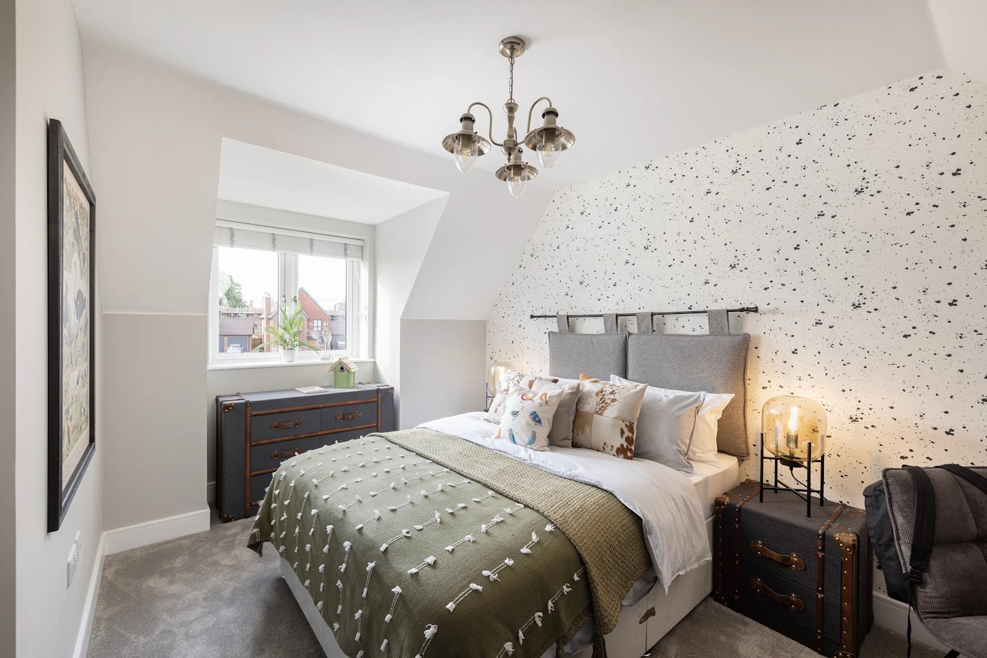 Cozy, contemporary bedroom featuring a double bed with a grey upholstered headboard, layered with white and patterned pillows and a green blanket accented with white foliage motifs. A clear-glass lamp sits atop a sleek white nightstand on the right, while a charcoal-toned dresser with brass handles anchors the left side, decorated with a small plant. Behind the bed, a feature wall with black-and-white speckled wallpaper contrasts the otherwise soft white walls. The room is softly illuminated by natural light through white-blinded windows and a three-light ceiling chandelier. A grey carpet grounds the space, enhancing the calm, inviting atmosphere