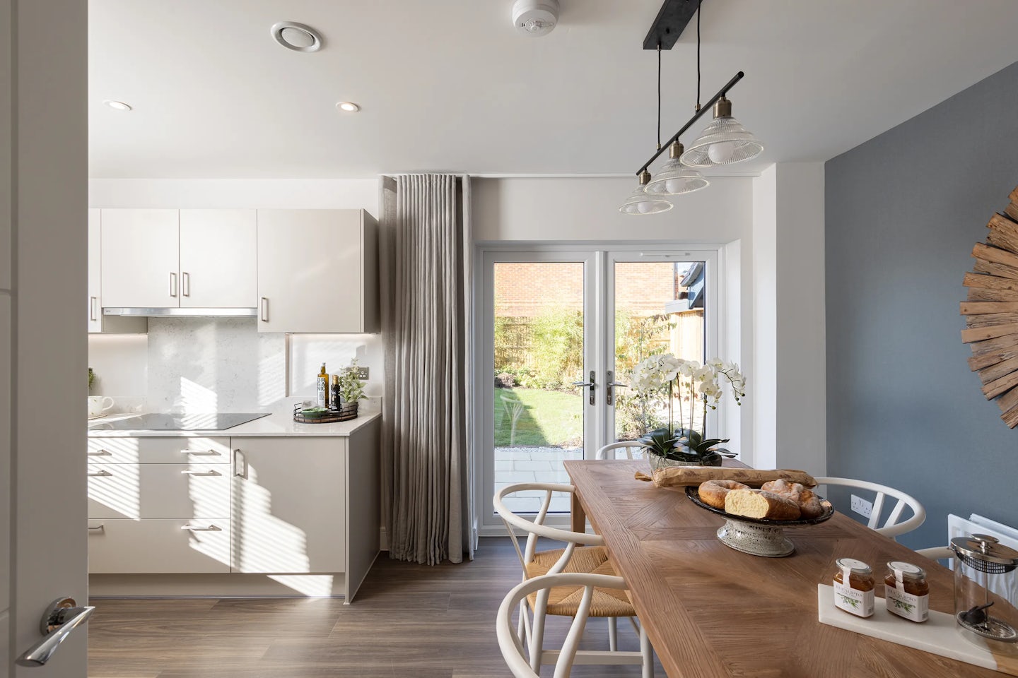Bright, contemporary kitchen and dining area featuring white cabinetry, a sleek backsplash, and a countertop adorned with glass bottles and a green potted plant. A large dining table sits center-stage, surrounded by white chairs with woven seats and decorated with assorted baked goods, glass jars, and a few small containers. Above, three pendant lights in matte black hang from the ceiling, balancing form and function. In the background, wide glass doors reveal a lush backyard with green grass and planting, bringing in natural light and extending the space visually. The soft palette of white, wood, and grey tones evokes a harmonious, welcoming atmosphere
