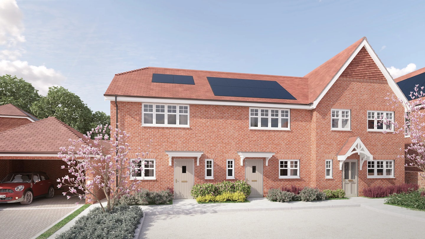 CGI of a modern red-brick townhouse complex with pitched tiled roofs and integrated solar panels. The building features three separate front doors, each framed by a small porch, suggesting individual units. Large first-floor windows and smaller ground-floor windows create a balanced façade. A red car is parked beneath a covered driveway on the left, while well-kept shrubs and flowering plants enhance the curb appeal. The scene unfolds on a clear day with a blue sky and light clouds, framed by surrounding trees and garden foliage to evoke a calm suburban atmosphere
