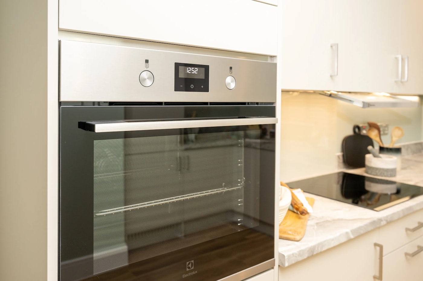 Modern kitchen featuring a built-in Electrolux oven with a digital clock reading “12:52,” framed by sleek white cabinetry with silver handles. The oven’s glass door reflects ambient light, while a nearby induction cooktop holds a saucepan. On the adjacent white countertop, a wooden cutting board displays a sliced loaf of bread and a knife beside neatly arranged utensils, evoking a moment of everyday preparation. The light-toned backsplash and clean layout enhance the contemporary, functional aesthetic.
