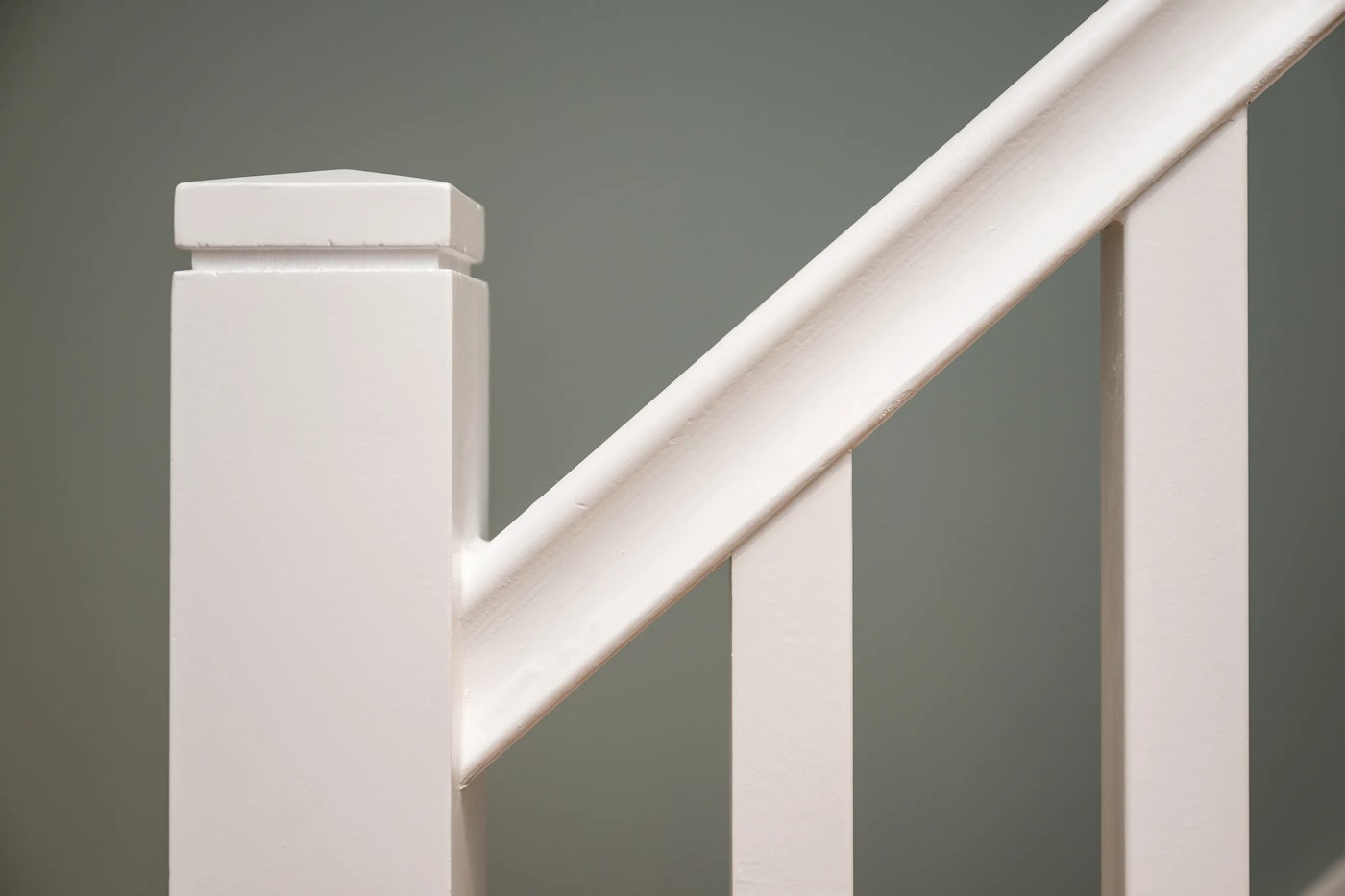 Close-up view of a white-painted staircase railing set against a muted green wall, featuring a square newel post with a capped top, angled handrail, and evenly spaced vertical balusters. A subtle circular mark sits at the centre of the handrail, drawing gentle visual focus. The clean lines and soft tones offer a quiet, orderly atmosphere with hints of architectural detail and domestic simplicity.