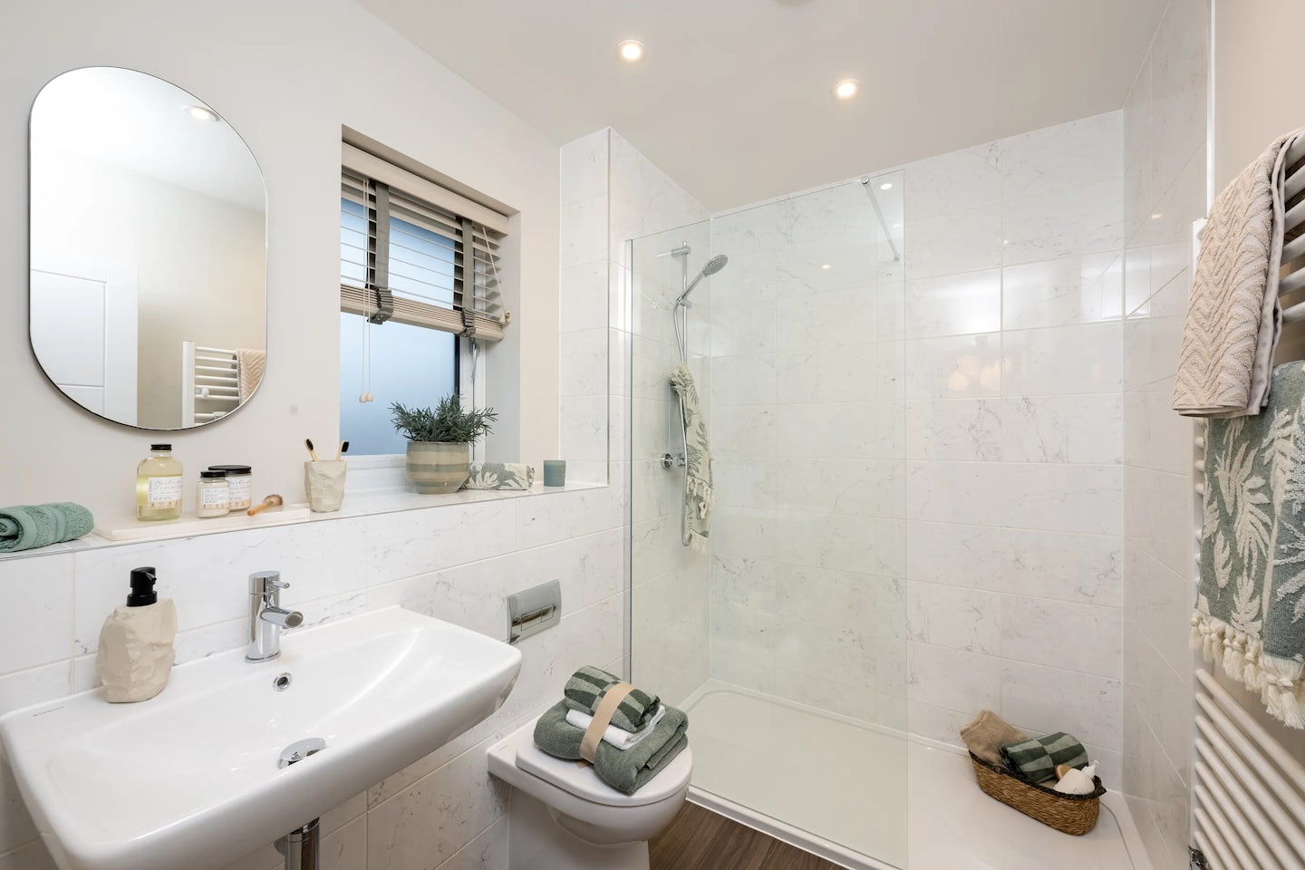 Bright, modern bathroom featuring a white sink with silver faucet, rectangular mirror, and window with white blinds above. A toilet sits nearby with neatly folded towels placed atop, while additional towels rest in a woven basket on the tiled floor. The countertop includes a soap dispenser, toothbrush holder, jar, and a small potted plant, adding personal charm. On the right, a large glass-enclosed shower with chrome fixtures completes the clean, minimalist design. White floor and wall tiles enhance the fresh, airy atmosphere