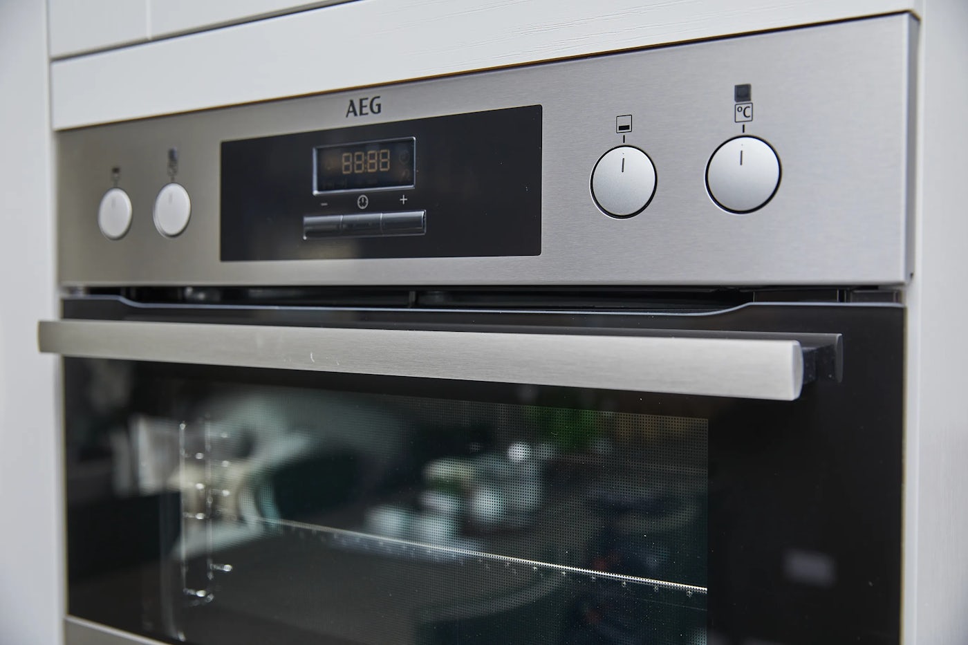 Close-up of a built-in AEG oven with a brushed stainless steel finish, featuring a digital display showing “8888” and two silver control knobs—one with a lightbulb icon and the other with a fan symbol beneath a “C.” The oven door is slightly ajar, revealing a wire rack inside. The clean lines and reflective surfaces evoke a sleek, modern aesthetic focused on practicality and understated design