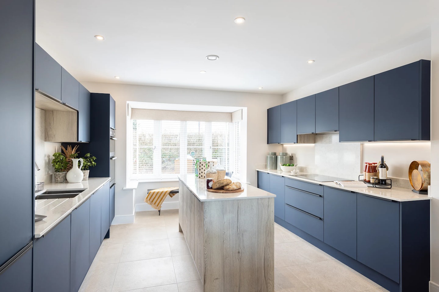 Sleek, modern kitchen featuring blue handleless cabinetry and light countertops, accented by a warm wood-finish island. A loaf of bread and kitchen essentials rest atop the island, adding lived-in charm. Recessed ceiling lights and a large window with white blinds provide ample illumination, enhancing the airy feel. On the counters, a coffee machine, cutting board, and potted plants suggest daily practicality. The tiled pale flooring complements the clean lines and balanced palette
