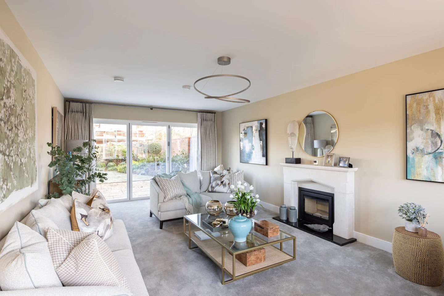 Elegant, light-filled living room featuring two cream sofas with plush cushions arranged around a rectangular glass coffee table with a gold frame. The table holds decor items including a blue vase, candle, and a small plant. A soft grey carpet grounds the space, while a circular ceiling light adds modern flair. Against the right wall, a white fireplace with a sculptural decor piece and a round mirror above serves as a focal point. Two abstract artworks flank the fireplace on pale beige walls, enhancing the refined aesthetic. Sliding glass doors in the background open to a lush garden