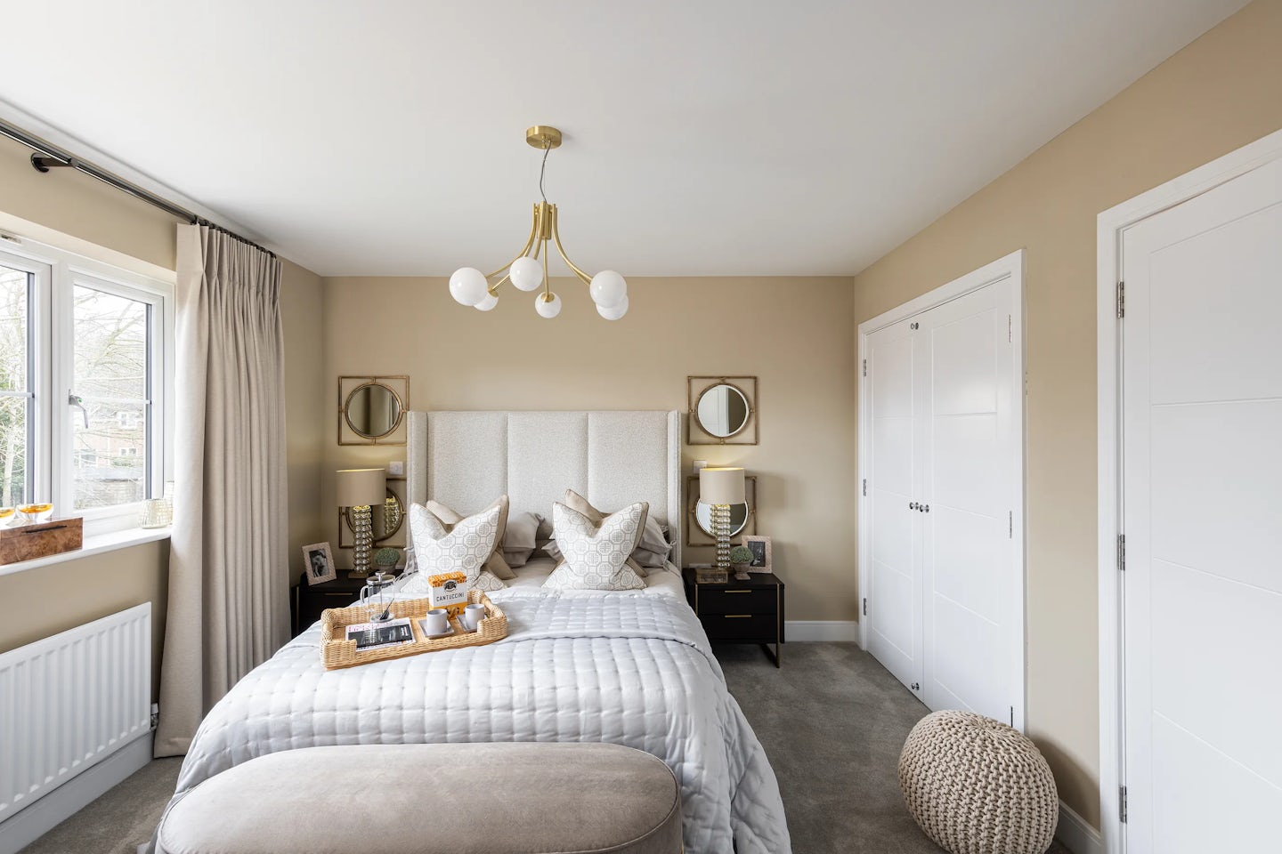 Elegant bedroom styled in soft neutrals, featuring a large bed with a padded beige headboard, crisp white linens, and an assortment of pillows. A wooden tray rests on the duvet, offering a breakfast scene with a croissant, orange juice, and coffee. Matching nightstands flank the bed, each topped with a lamp and curated decor. A modern chandelier with four spherical bulbs hangs above, adding sculptural flair. Beige walls and a grey carpet set a muted backdrop, while beige curtains soften the window light. To the right, a double-door white closet and a round knitted pouf