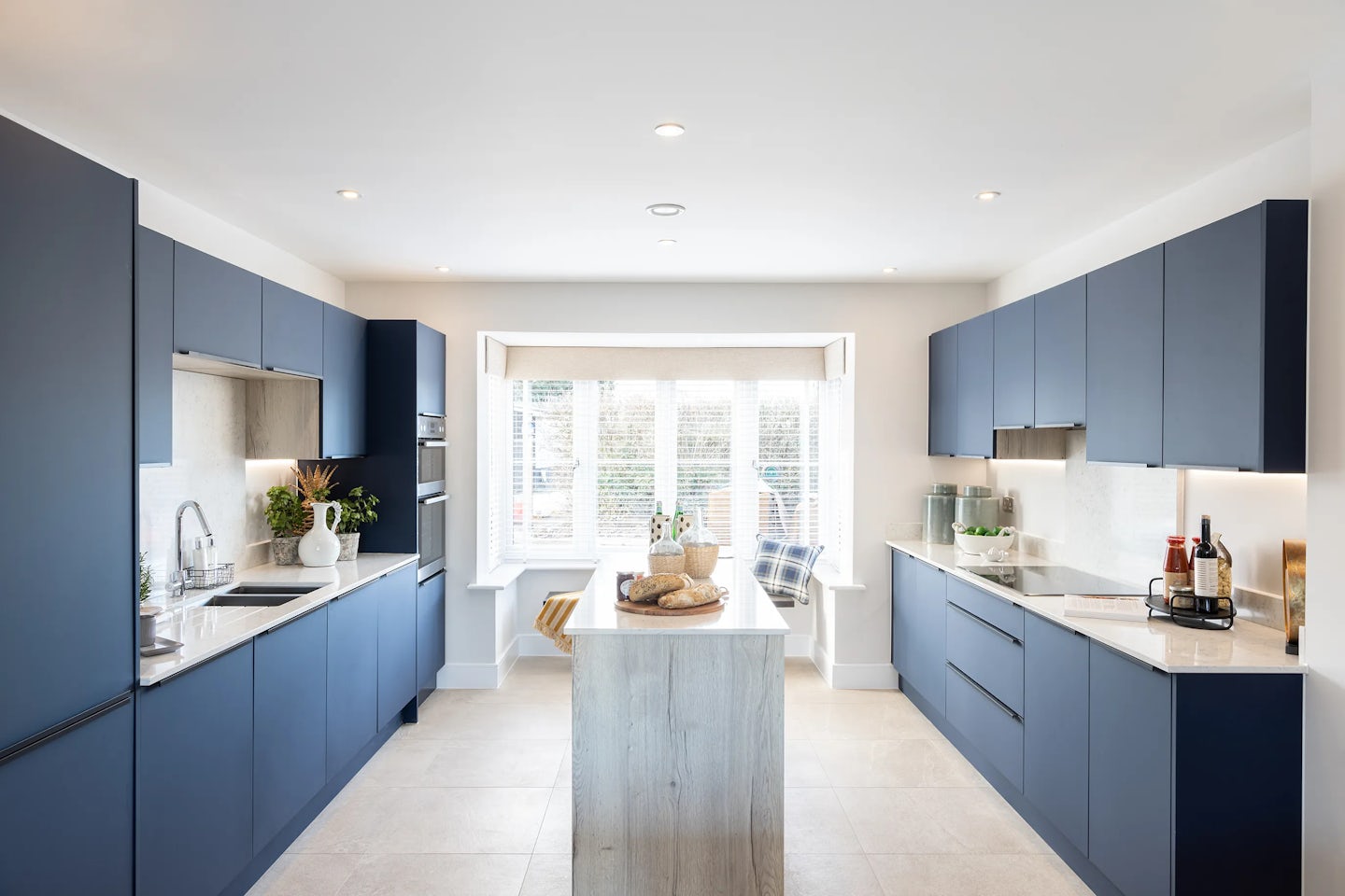 Contemporary galley kitchen featuring deep blue cabinetry with sleek silver handles, white countertops, and a warm wood-toned central island. Built-in appliances—including an oven and microwave—integrate seamlessly on the left, while a stovetop anchors the right. A coffee machine, cutting board, and potted plants personalize the space. The far end showcases a cozy window seat dressed with cushions, positioned beneath large windows that let in soft natural light. Recessed lighting above and pale flooring below enhance the room’s bright, balanced atmosphere