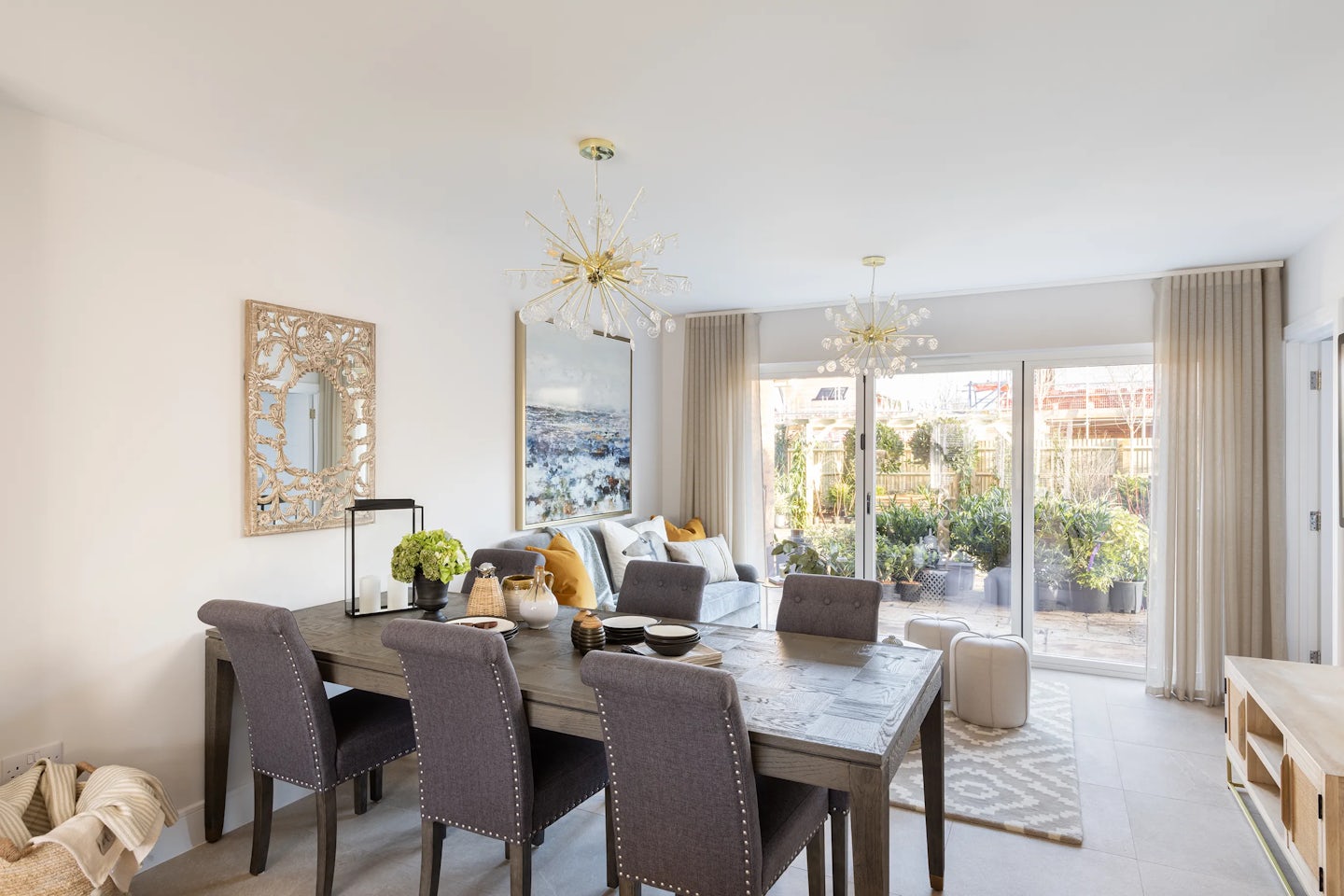 Spacious, light-filled living and dining area featuring a wooden dining table with six upholstered chairs, styled with a floral centerpiece and candle set. A large decorative mirror anchors the dining wall, enhancing depth and brightness. The living space includes a neutral-toned sofa layered with cushions, set beneath an abstract seascape painting. A rectangular coffee table displays vases and a small plant, complementing the relaxed elegance. Two chandeliers hang overhead, adding symmetry and sparkle.Full-height glass doors open to a lush garden with varied greenery, extending the room’s serene flow into the outdoors.