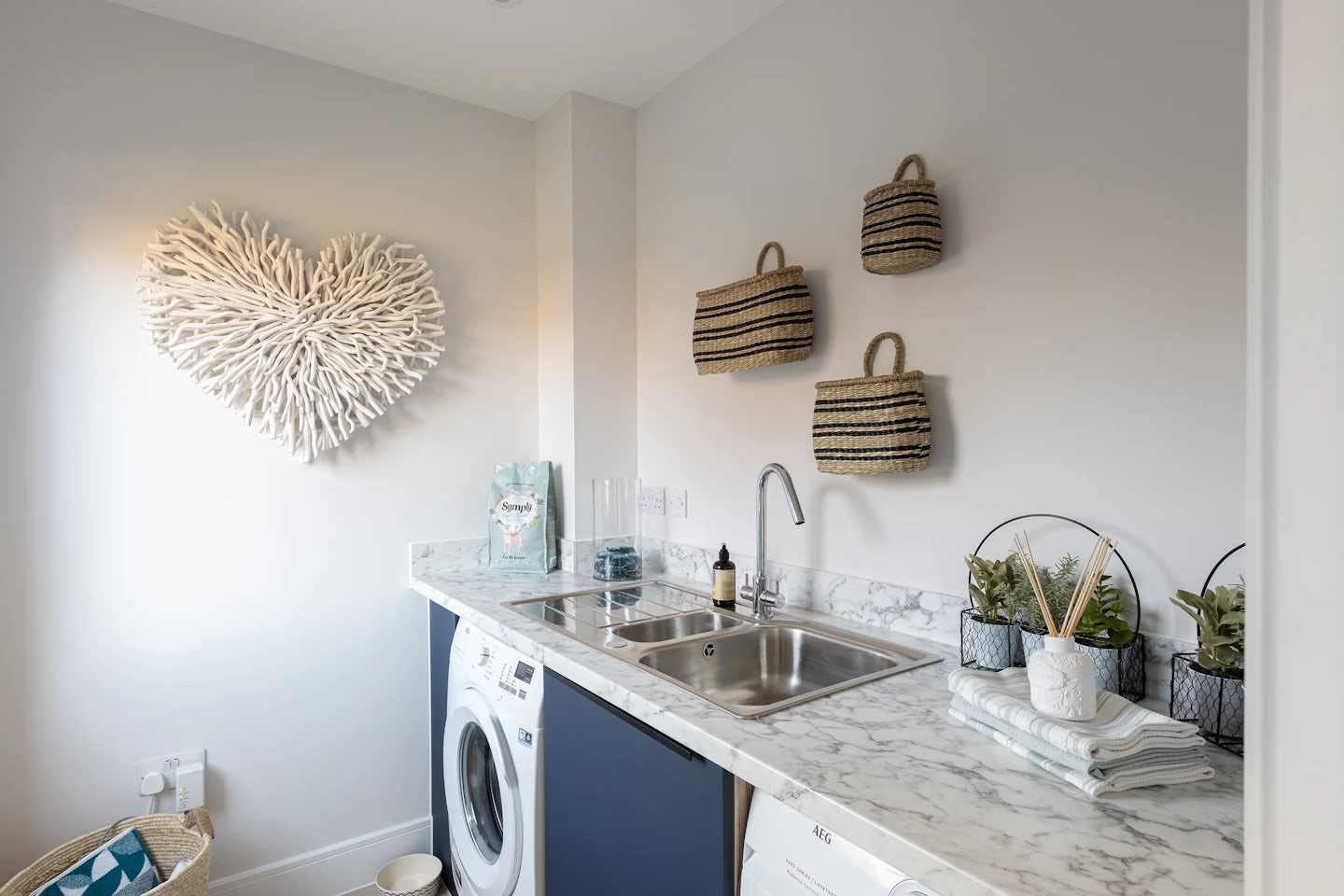 Well-organized, contemporary laundry room featuring a white AEG washing machine and dryer installed beneath a marble countertop. To the right, a stainless steel sink with a chrome faucet sits beside a bottle of hand soap, a glass container with blue items, and a wire basket holding small potted plants. Folded towels rest neatly atop the counter, and a trio of woven wall baskets hang above the sink. On the left, a heart-shaped wall decor crafted from white coral-like material adds texture and charm, while a wicker basket with a blue-and-white pillow anchors the floor space.