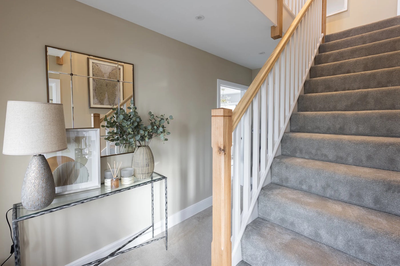 Inviting hallway interior featuring a soft grey-carpeted staircase with white railings and warm wooden posts. A black metal-framed console table with a glass top sits to the left, styled with a textured lamp, framed photo, candle, reed diffuser, and a vase of green foliage. Above, a large grid-style mirror reflects the neutral-toned space, adding depth and symmetry. Light beige walls and matching carpet throughout offer a cohesive, calm aesthetic.