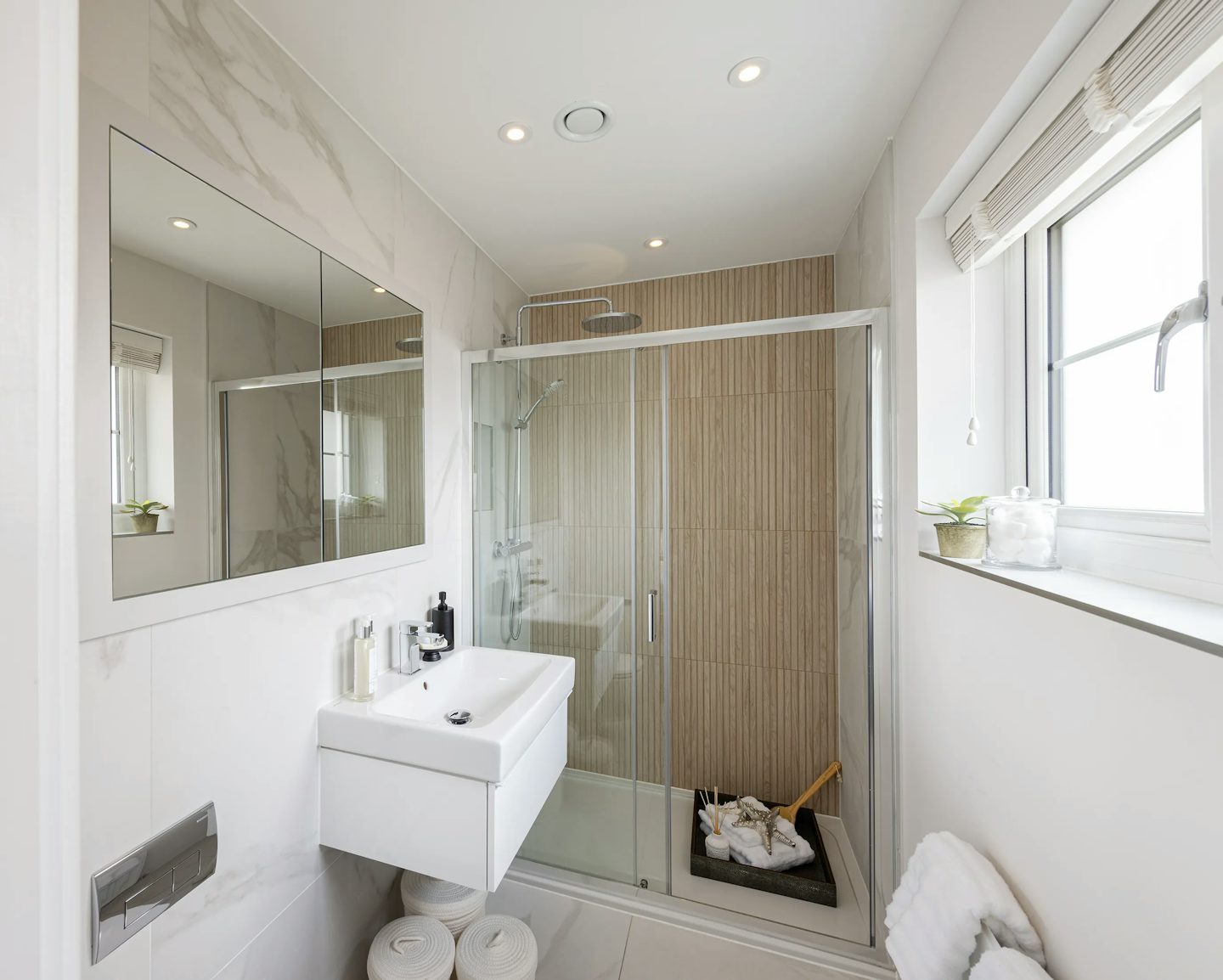 Modern, compact bathroom with a clean, minimalist layout. A glass-enclosed shower with a wood-textured back wall adds warmth and style, while a white sink beneath a rectangular mirror anchors the right side. The countertop holds everyday toiletries and a sleek soap dispenser, offering lived-in practicality. Under the sink, neatly rolled towels enhance the sense of order. Soft light filters through a window fitted with white blinds, its sill hosting a small potted plant for a touch of greenery. Pale tiled flooring and recessed ceiling lights contribute to the bright, uncluttered ambiance