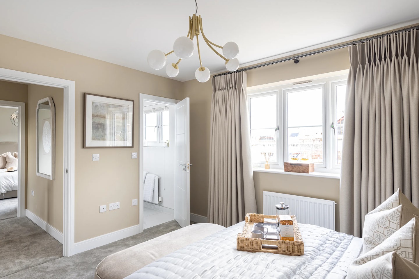 Tranquil bedroom with soft beige walls and a bed dressed in white and beige linens, accented by patterned pillows. A woven tray with a teapot and cups rests atop the duvet, enhancing the scene’s lived-in warmth. A modern chandelier of spherical bulbs hangs centrally, adding sculptural charm. To the left, an open door reveals a bright bathroom with white walls and a neatly hung towel. Another door leads to an adjacent space, possibly another bedroom. The large window—framed by beige curtains—offers a view of rooftops and sky