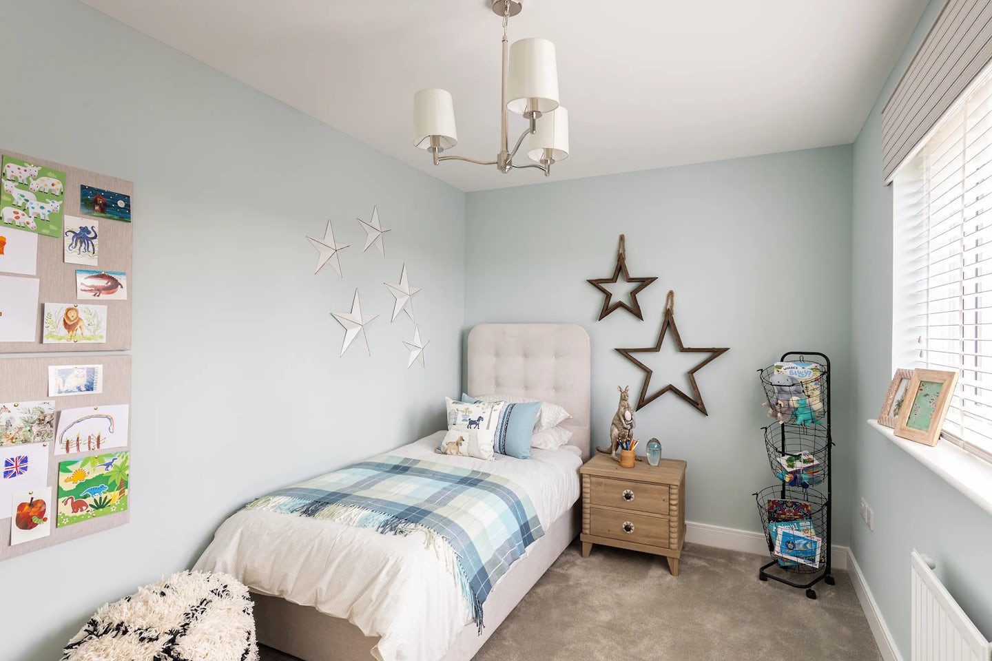 Cozy child’s bedroom featuring a single bed dressed in a blue-and-white plaid comforter with layered pillows, including a decorative star-print cushion. Above the bed, five white star-shaped wall hangings add playful symmetry. A wooden nightstand to the right holds a small lamp, a figurine, and glass decor, while two large star decorations above it echo the room’s celestial motif. A metal shelving rack beside the nightstand displays books and toys, contributing to the organized yet inviting atmosphere. On the opposite wall, a bulletin board showcases vibrant child-made drawings of animals, nature, and flags above a plush white rug.