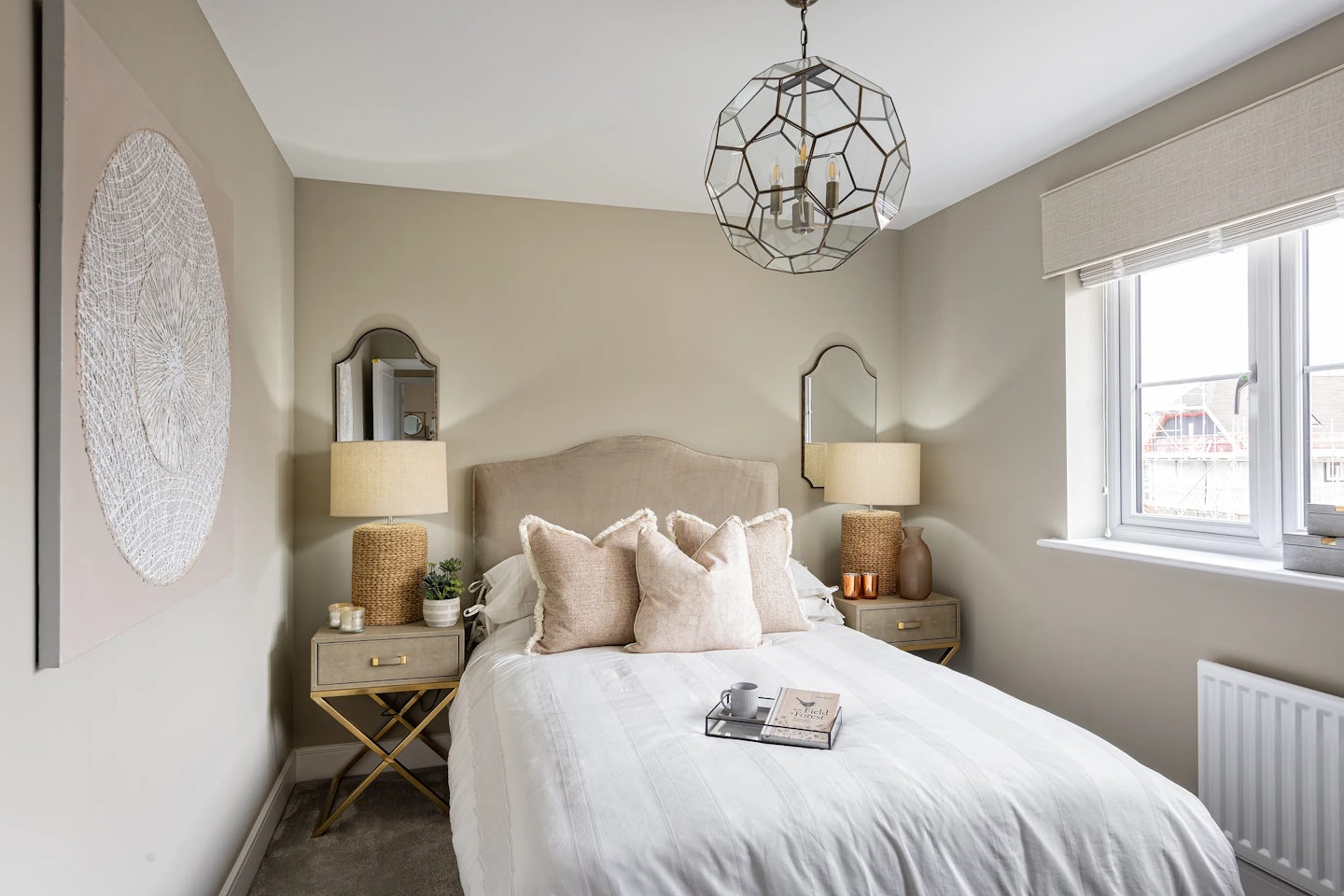 Refined bedroom styled in soft neutrals, featuring a bed with a beige upholstered headboard, crisp white bedding, and assorted beige and white pillows. A tray with a cup and open book rests atop the duvet, adding lived-in warmth. Beside the bed, two nightstands each hold a lamp, small plant, and ceramic accent. Above, a geometric chandelier provides sculptural flair, while circular wall art and framed mirrors enhance symmetry