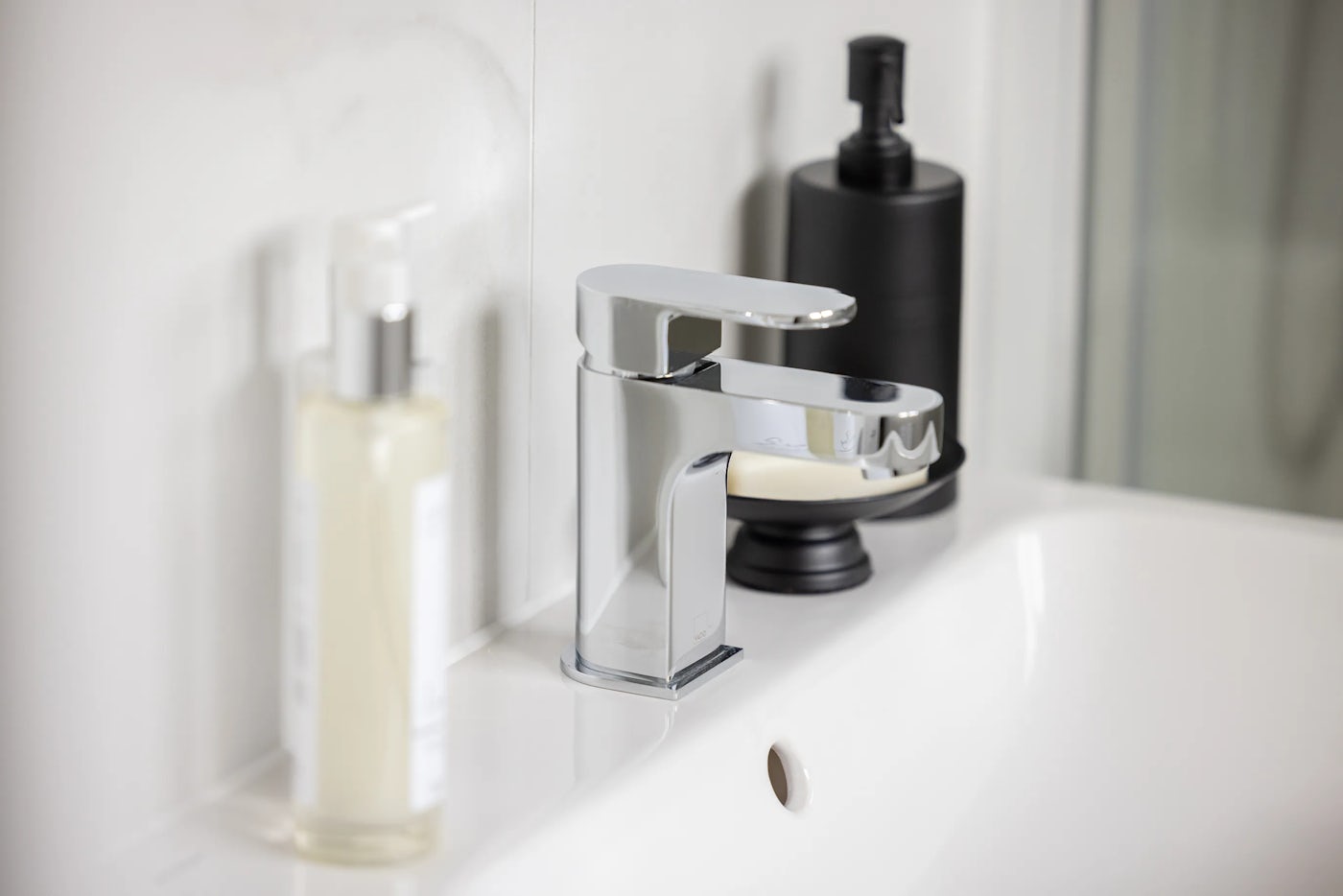 Close-up of a white bathroom sink with a curved chrome faucet positioned centrally. On the right sits a matte black soap dispenser with a metallic pump, while on the left a translucent bottle with a silver pump top adds visual contrast. The drain hole is clearly visible in the foreground, and the surrounding surfaces are clean, minimal, and gently reflective, suggesting modernity and functional elegance