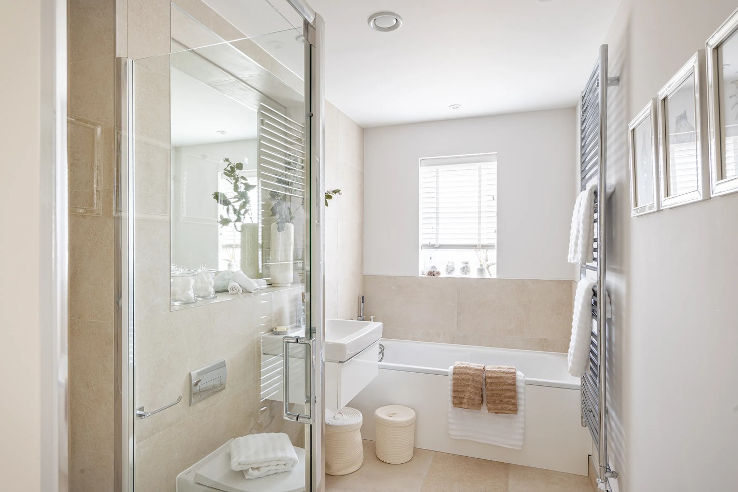 Modern, light-filled bathroom featuring a glass-enclosed shower on the left and a beige-tiled bathtub with a neatly draped white towel. A white vanity unit with twin cylindrical wicker baskets sits beneath a wall-mounted sink and large mirror, while a nearby window with blinds allows soft natural light to pour in. On the right wall, a towel rack holds stacked white towels, framed by three decorative wall prints above.