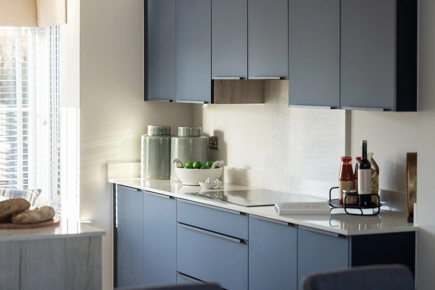 Modern kitchen with navy blue cabinetry and white countertops, styled with a mix of fresh and functional touches. Two large green jars and a white bowl filled with green apples sit centrally, while a loaf of bread on a wooden cutting board adds rustic warmth. A black wire rack holds olive oil bottles, sauces, and seasonings, suggesting everyday culinary readiness. Soft natural light filters through a window with white blinds above the sink, illuminating the clean lines and calm palette of the space.