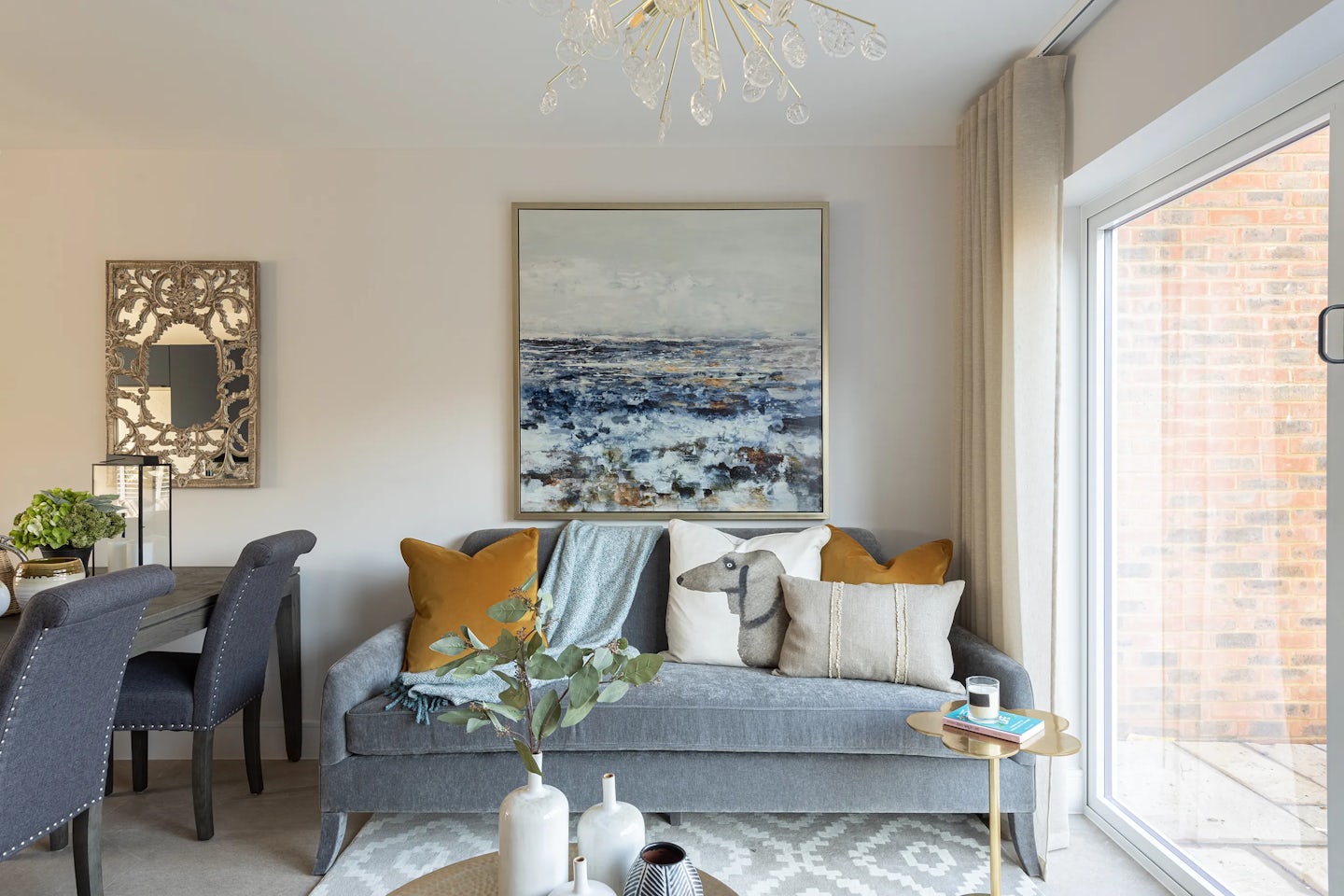 Well-lit living room blending modern comfort with artistic flair. A grey sofa adorned with mustard and patterned cushions sits centrally beneath a large abstract seascape painting. To the left, a dining area features a wooden table and upholstered chairs, accompanied by a decorative wall mirror. A small round side table beside the sofa holds a lit candle and an open book, while the rectangular coffee table displays a plant and two vases, enhancing the room’s relaxed elegance.
