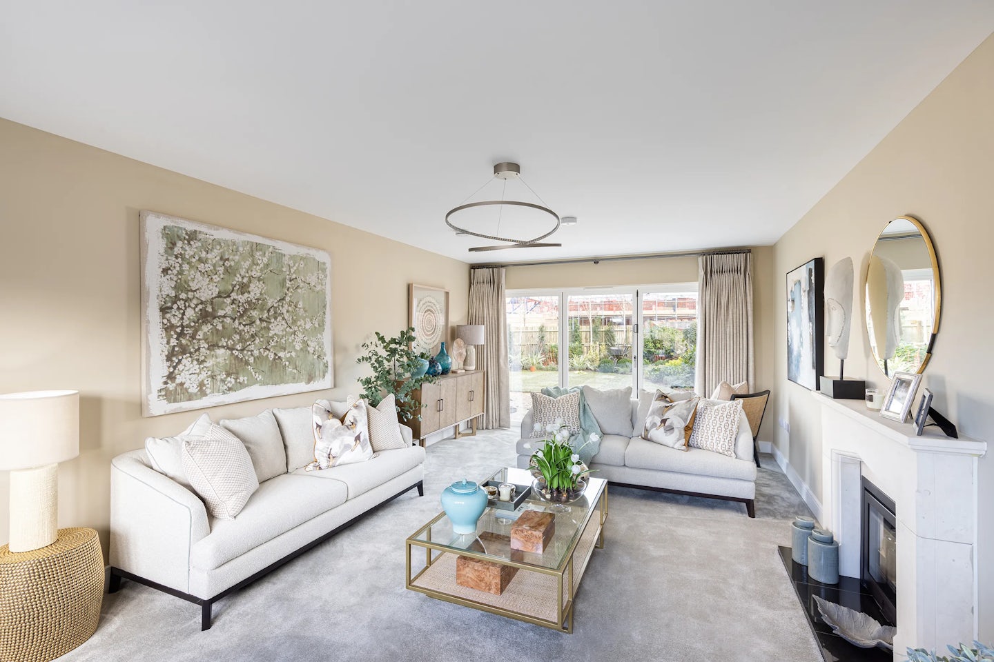 Refined living room styled in soft neutrals and natural textures. Two light-colored sofas with patterned cushions face a rectangular glass coffee table, which holds a blue vase, stack of books, and a small plant. A fireplace anchors the right wall, topped with decorative vases, a lamp, and a round mirror that adds symmetry and light. To the left, a large abstract artwork lends visual depth. A spacious window dressed with sheer curtains fills the room with daylight, softening the beige walls and grey carpet. The overall ambiance combines comfort, spaciousness, and understated elegance