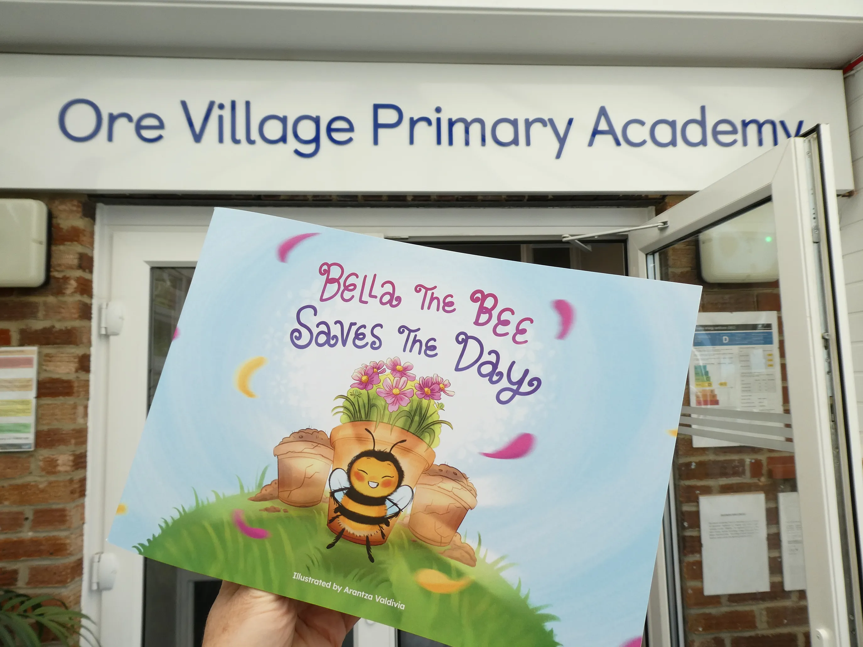 A person standing outside Ore Village Primary Academy, holding a children’s book titled “Bella The Bee Saves The Day.” The cover features a cartoon bee with a superhero cape, surrounded by flowers and petals on a grassy field. The book is illustrated by Antonia Valerio. The academy entrance, with open doors and posted notices, is visible in the background.