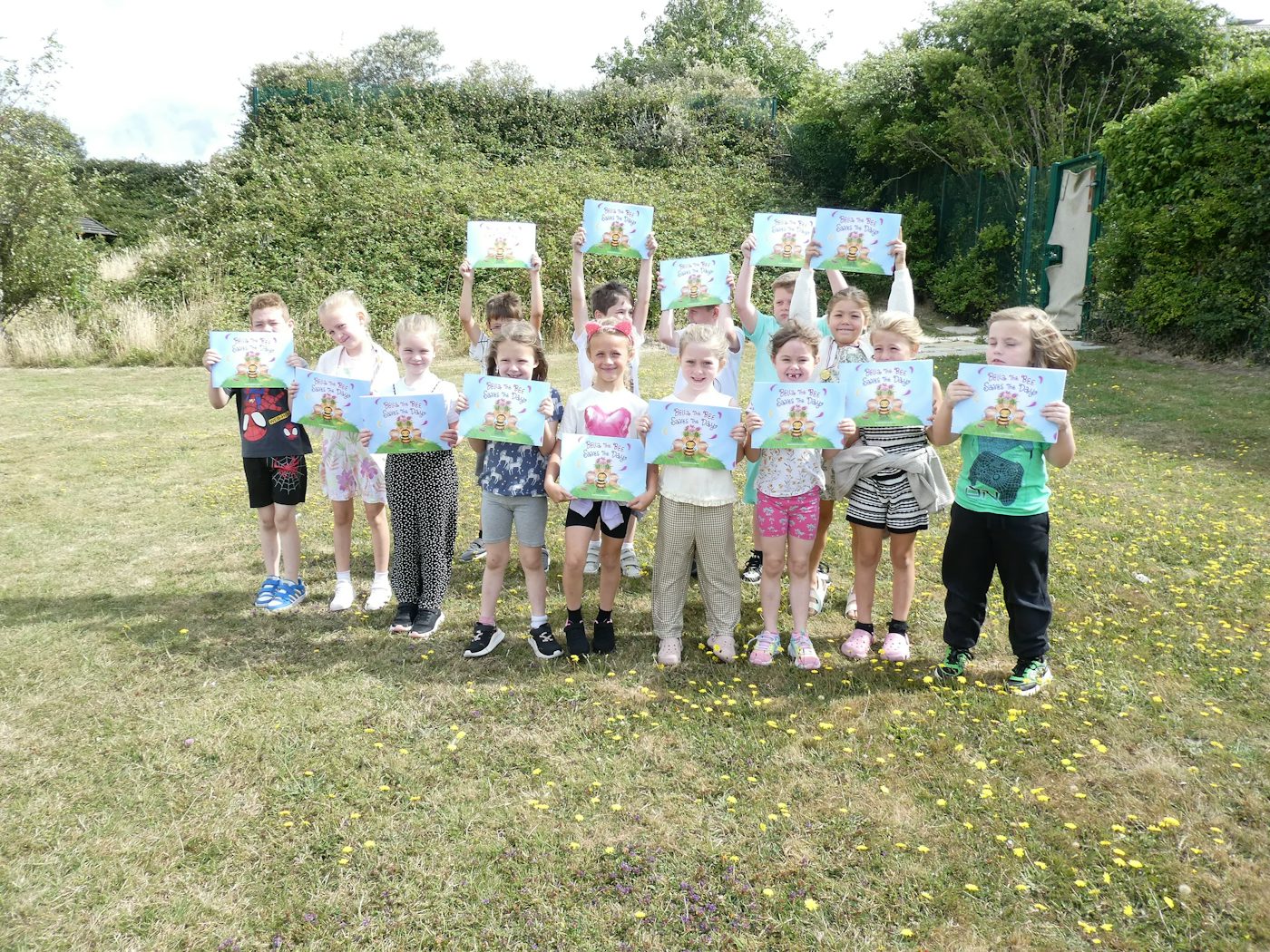 A group of children outdoors on a grassy area, smiling and holding up colorful certificates or cartoon-themed artwork. They’re arranged in two rows, with some kneeling in front and others standing behind. Trees and part of a building appear in the background, adding to the summery, celebratory atmosphere.