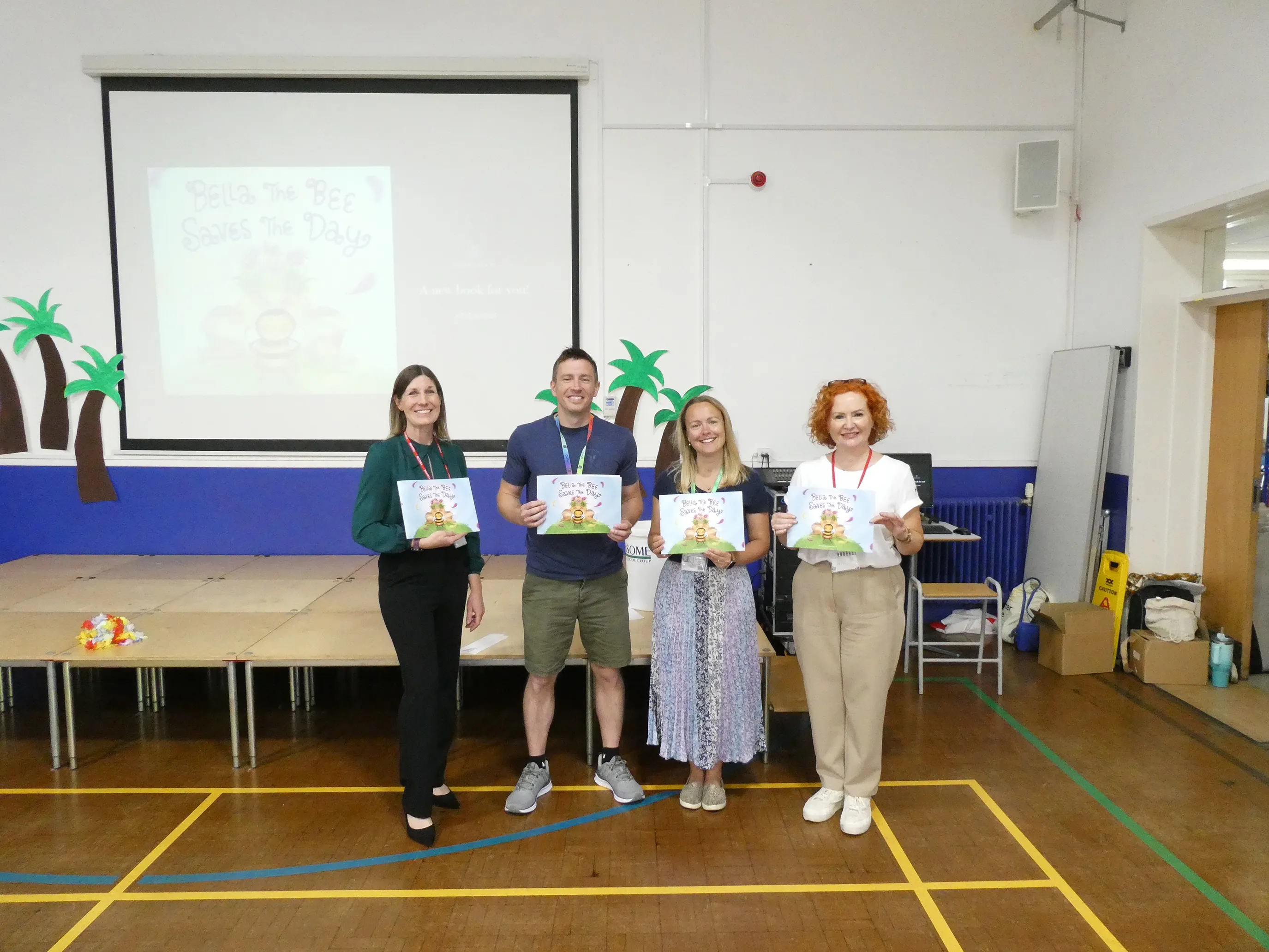 Four individuals standing in a decorated room, each holding a copy of the children’s book “Bella the Bee Saves the Day.” A projector screen behind them displays the book cover, showing a cartoon bee in a superhero cape. The room features tropical-themed decor, including palm trees, with tables and items arranged for a presentation or event. All four wear lanyards, suggesting they’re part of an official launch or workshop