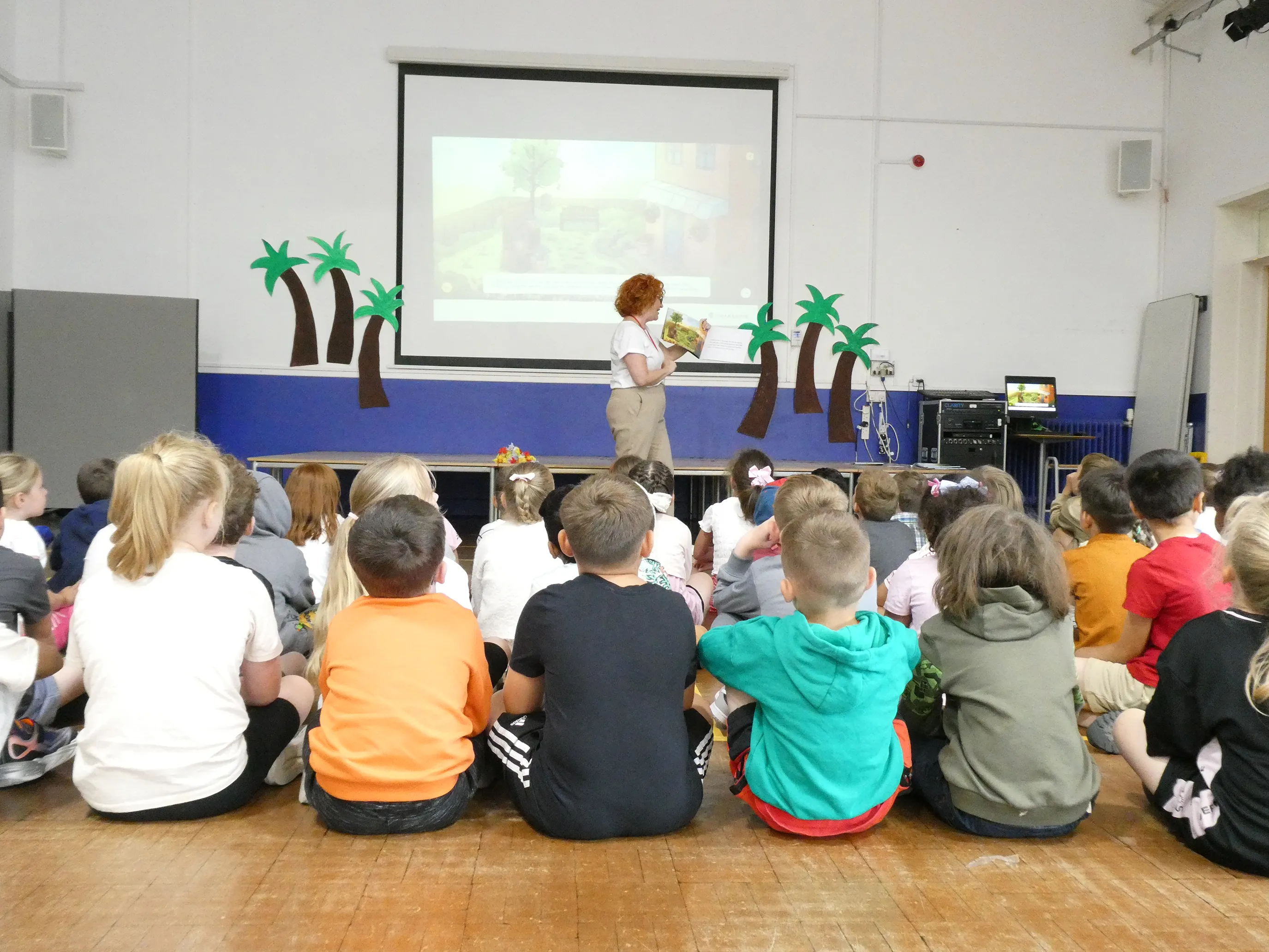 A person stands in front of a group of seated children, presenting a storybook titled “Bella the Bee Saves the Day.” Behind them, a screen displays the book’s cover, showing a cartoon bee with a superhero cape. The classroom is decorated with tropical palm tree cutouts, adding a playful atmosphere. A computer and projector setup is visible beside the screen, suggesting an interactive storytelling session.
