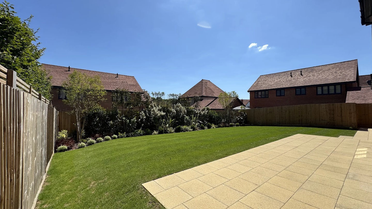 A sunny backyard featuring a well-kept lawn, a paved patio area, and a wooden fence enclosing the space. Flowering shrubs and young trees line the perimeter, adding seasonal greenery. In the distance, neighbouring houses with red-tiled roofs peek over the fence under a clear blue sky, creating a warm, suburban atmosphere