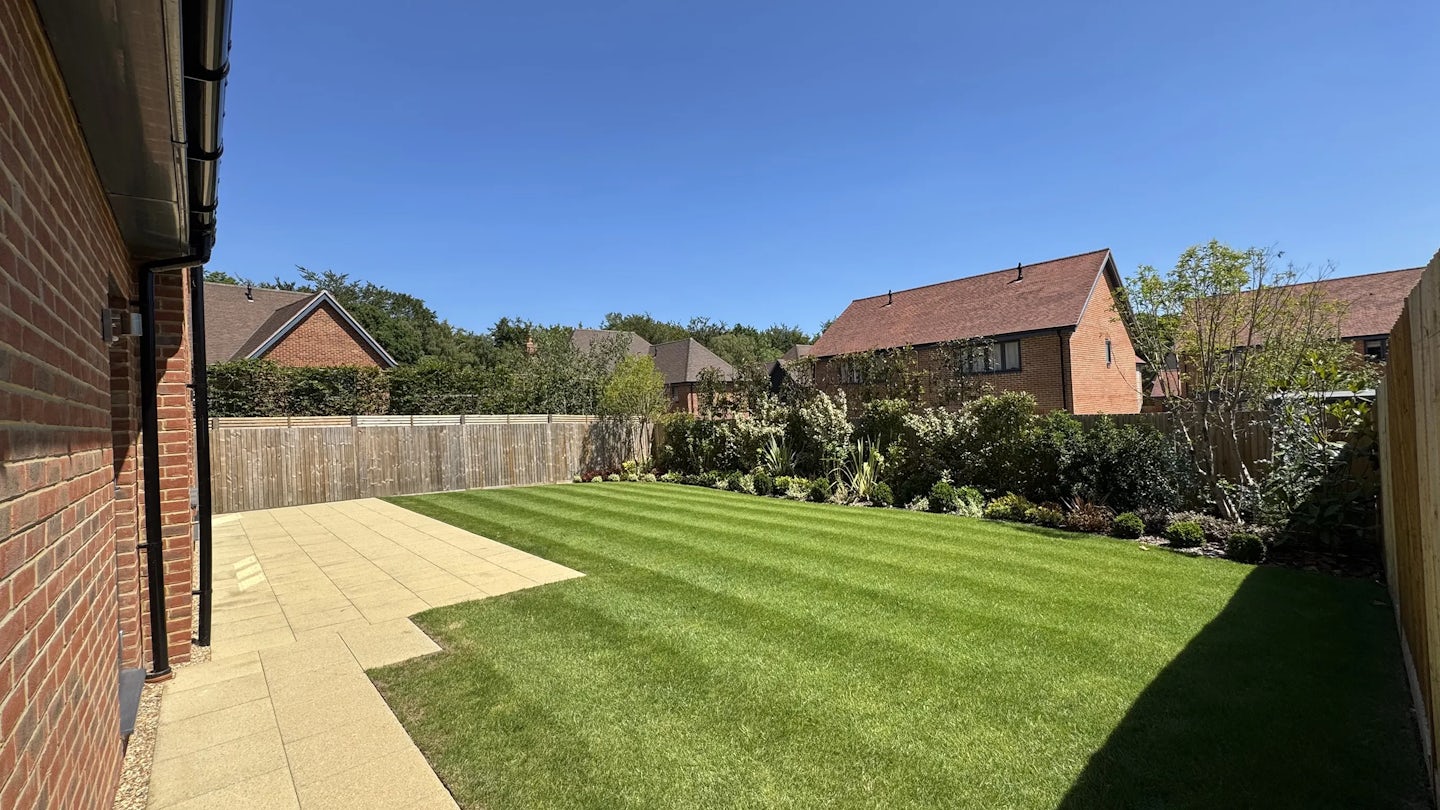 A neatly maintained backyard with a freshly mowed lawn showing alternating green stripes. The paved patio borders a red-brick house, and a wooden fence encloses the garden. Shrubs and leafy plants line the edges, with neighboring red-brick homes visible beyond. The scene is bathed in warm sunlight under a clear blue sky, giving a calm, summery feel