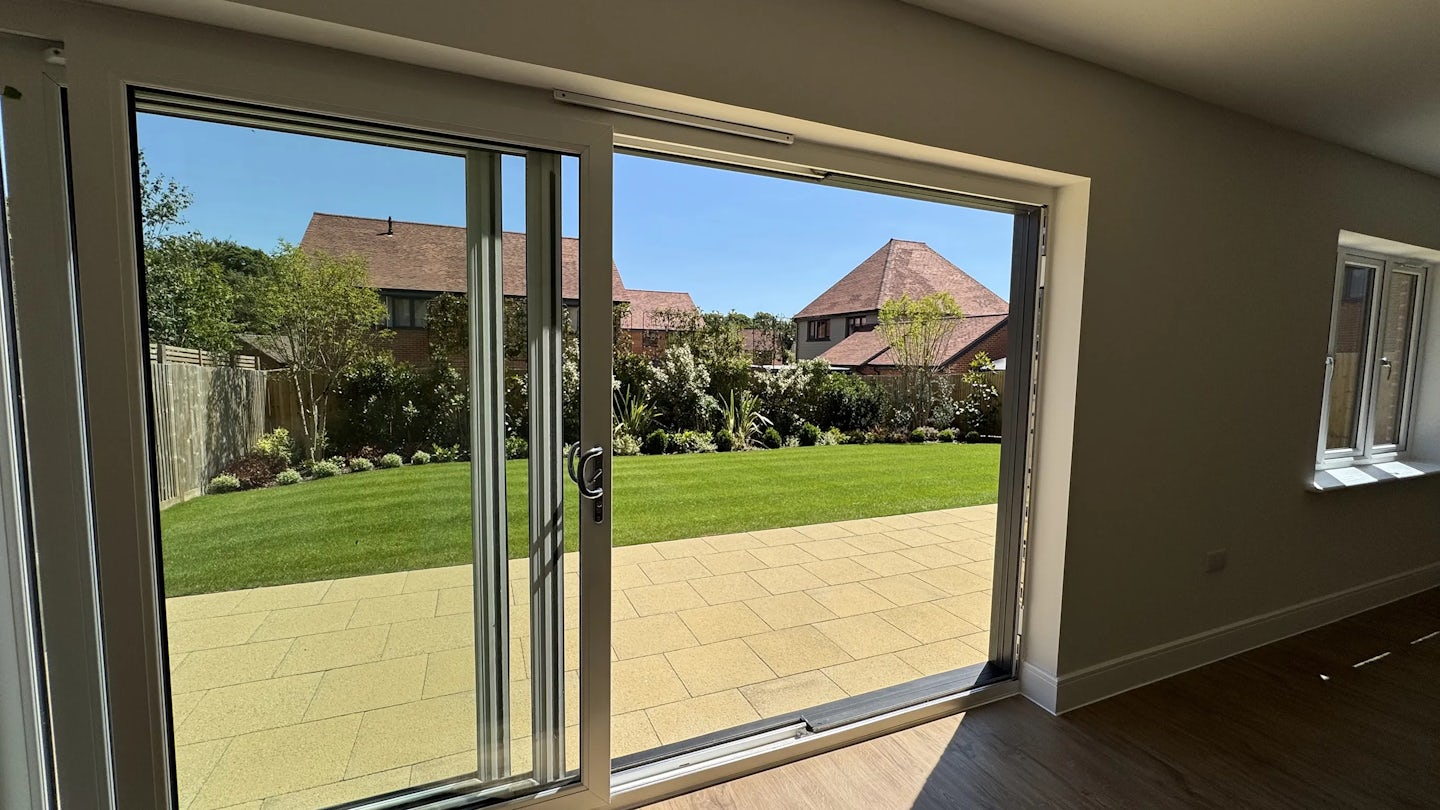 View from inside a room with wooden flooring, looking out through a large sliding glass door onto a sunny garden. The garden features a neatly trimmed lawn, shrubs, and flowering plants, with neighboring houses and leafy trees visible beyond. A window on the right and a clear blue sky add to the calm, summery feel
