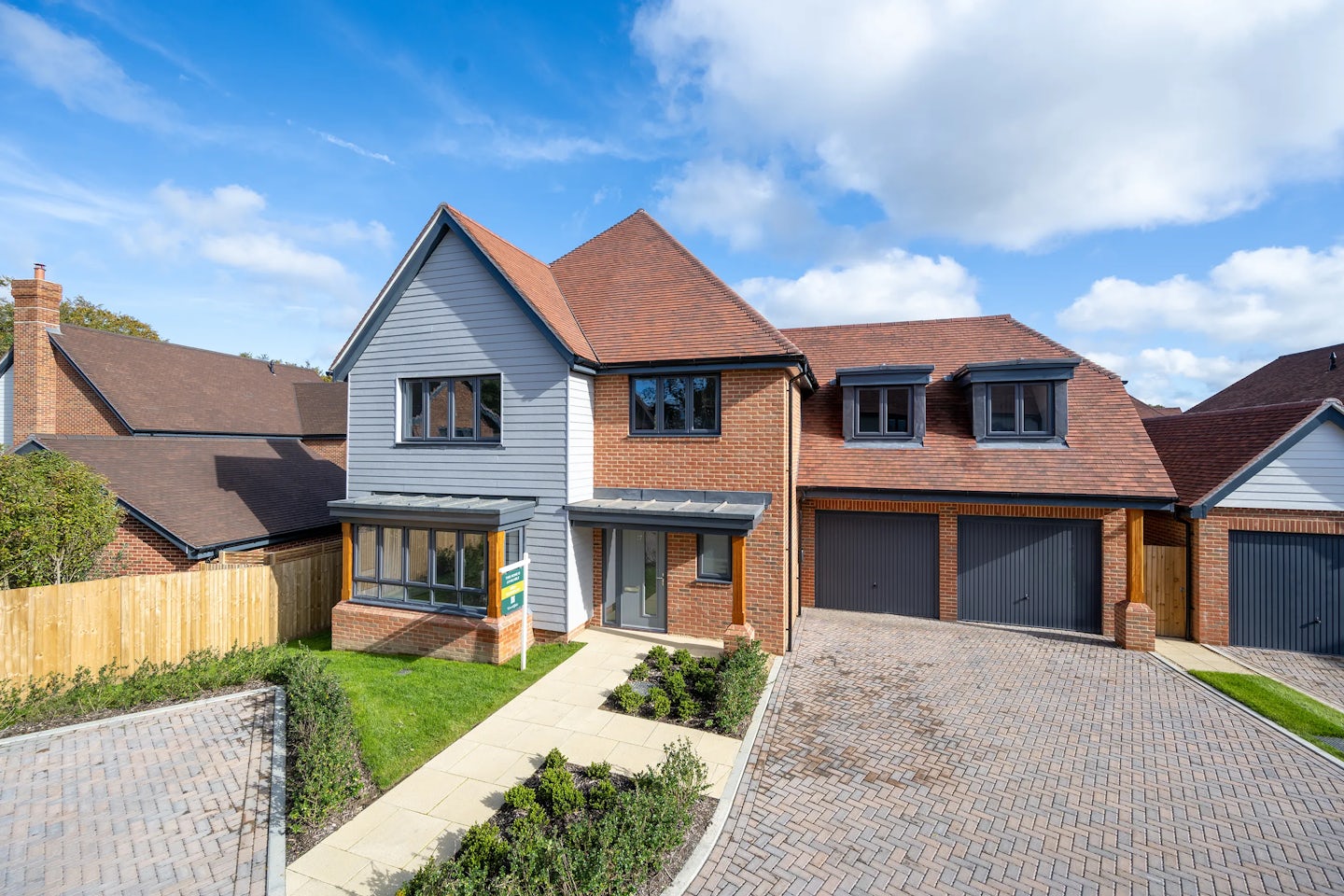 A modern two-story house with a red-tiled roof and a mix of brick and grey cladding on the exterior. The home features a double garage with grey doors, a paved driveway made of interlocking bricks, and a small front lawn bordered by a walkway leading to the front door. Large windows, including a bay window on the ground floor and dormer windows on the upper level, enhance the façade. A partly cloudy sky overhead adds to the pleasant, suburban atmosphere