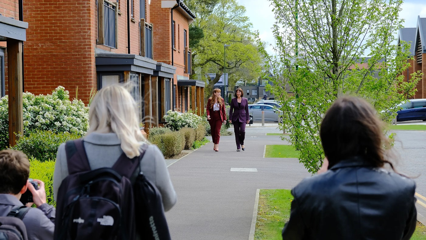 Several people are gathered on a residential street, with three individuals in the foreground photographing or filming two women walking nearby. The women, dressed in burgundy and dark purple formal outfits, appear to be the focus of the shoot.