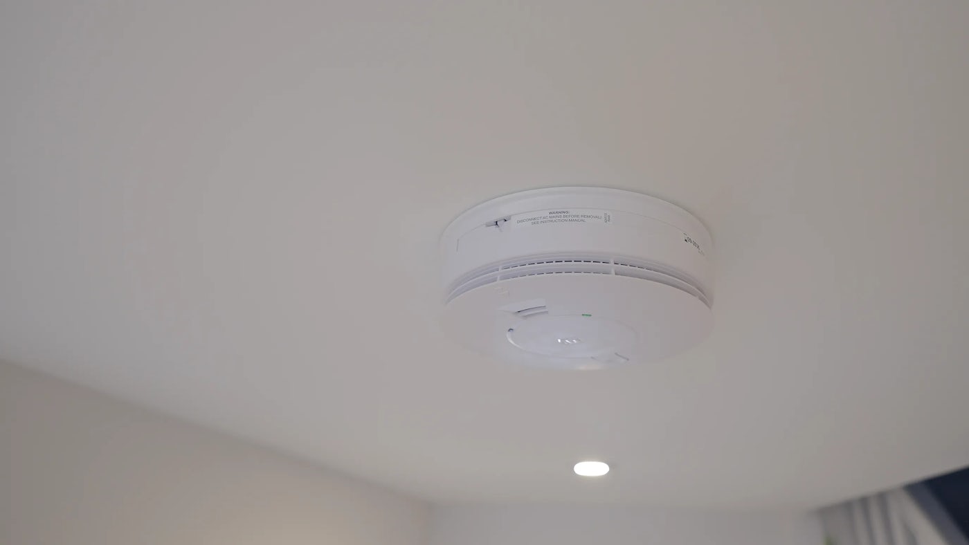 A close-up of a white smoke detector mounted on a ceiling. The device is round with vented edges and features a small green indicator light near the center.