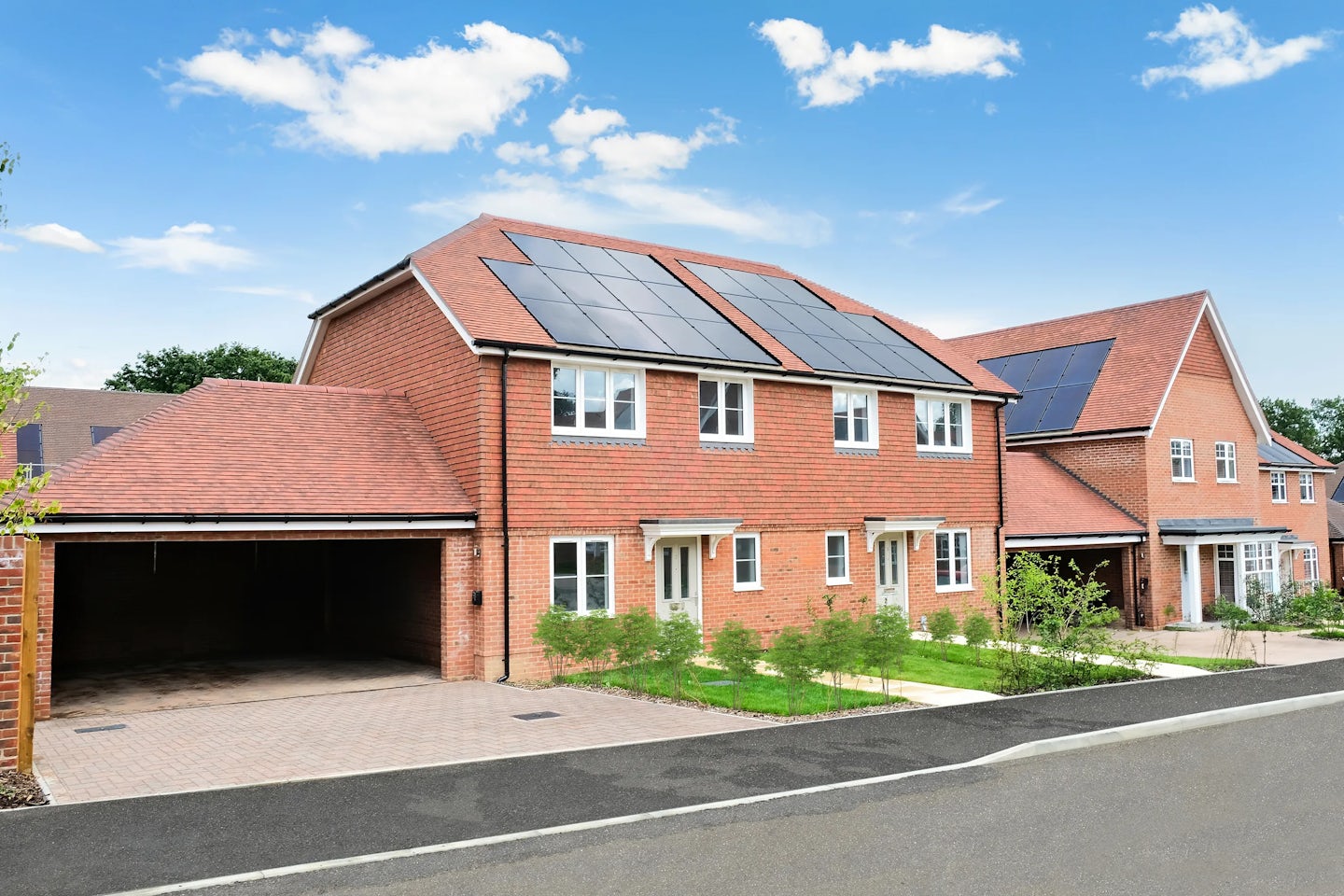 A row of modern brick houses in a well-kept neighborhood. The house in the foreground features solar panels on the roof, a driveway leading to a garage, and a small patch of greenery. The paved road runs along the front, and the sky is mostly clear with a few clouds, suggesting a bright, pleasant day.