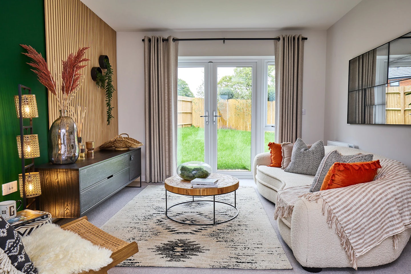 A stylish, light-filled living room with a white sofa accented by orange and gray cushions, facing a round glass-topped coffee table on a patterned rug. A large window and glass door open to a green backyard. The space features a wood-slatted wall with a green accent, a sideboard with decorative items, and a sculptural floor lamp. A grid-pattern mirror hangs on the opposite wall, adding depth to the cozy, modern setting.