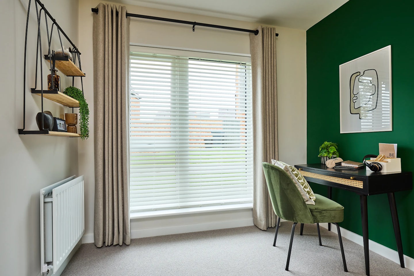 A stylish home office with a large window dressed in white blinds and beige curtains. A black desk with cane-front drawers sits against a green accent wall, paired with a green upholstered chair. The desk holds a plant, notebook, and headphones. Above the desk hangs a framed abstract artwork. A wall-mounted shelf to the left displays books, a small plant, and decorative items. The room combines natural textures with a modern aesthetic, creating a calm and productive workspace
