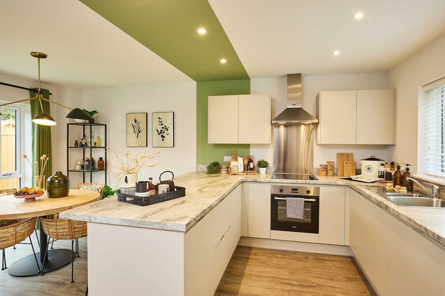 A bright, modern kitchen and dining space featuring a U-shaped marble countertop, white cabinets, and stainless steel appliances. A green accent wall adds contrast above the stove. The counter holds a tray with utensils, a potted plant, and a bowl of fruit. Adjacent is a round wooden dining table with wicker chairs beneath a hanging light fixture. Two framed artworks and a shelving unit with decorative items enhance the cozy, well-lit room, which receives natural light through a window with white blinds