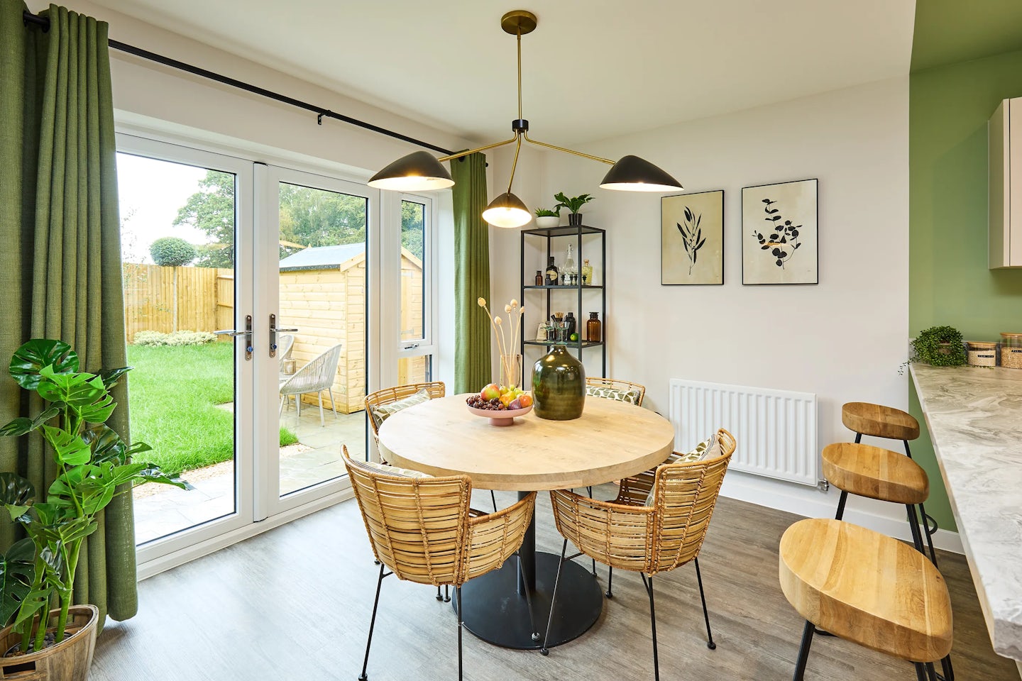 A cozy, modern dining area featuring a round wooden table with four wicker chairs beneath a trio of pendant lights. A green vase and fruit bowl sit atop the table. Green curtains frame glass doors that open to a garden with a wooden shed and outdoor seating. To the right, a kitchen counter with two wooden bar stools adds functionality. The space is accented by botanical prints, open shelving with plants and bottles, and light wood flooring, creating a warm, inviting atmosphere.