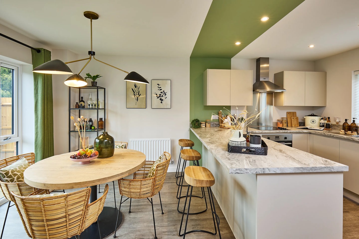 A bright, modern kitchen and dining space featuring a marble breakfast bar with two wooden stools, white cabinetry, and stainless steel appliances. A round wooden dining table with wicker chairs sits beneath a trio of pendant lights. Green curtains frame a window, complementing the green accent wall behind open shelves filled with plants and bottles. Two framed artworks hang above a white radiator, adding a personal touch to the well-lit, inviting room.