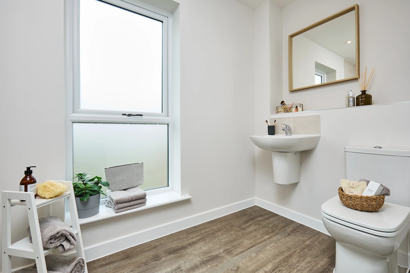 A modern bathroom with a white wall-mounted sink and chrome faucet beneath a large wooden-framed mirror. To the right, a white toilet holds a wicker basket filled with toiletries. A frosted glass window above the toilet lets in light, with folded towels and a small plant on the windowsill. A white shelving unit beside the window stores more towels and a brown pump bottle. The space features wood-look vinyl flooring and a clean, minimal aesthetic