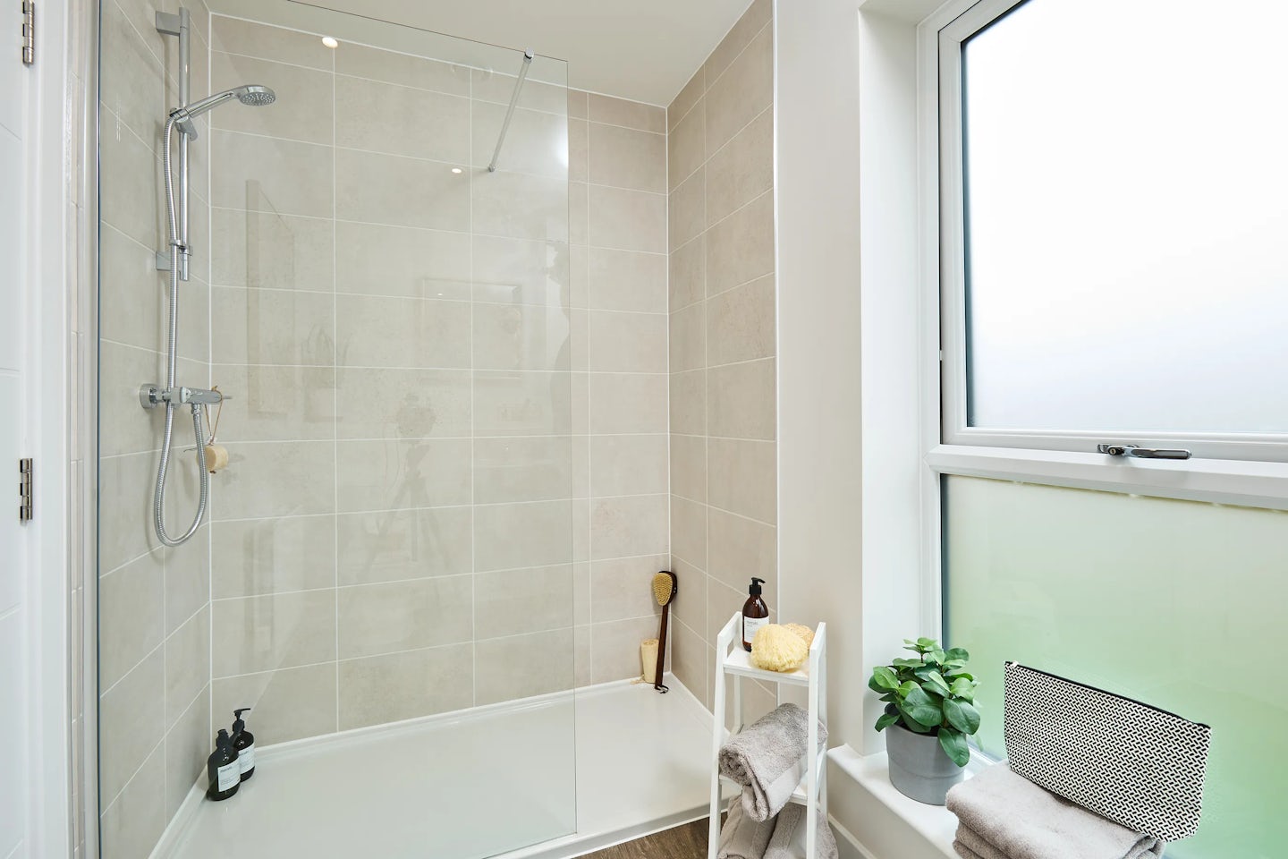 A modern bathroom with a walk-in shower enclosed by a glass partition and beige tiled walls. Inside the shower, a silver showerhead and soap holder are mounted above two black bottles on the floor. Outside the shower, a white shelf holds folded towels, a brown brush, a yellow sponge, and a bottle. A potted plant sits on the windowsill, with frosted glass on the lower half for privacy. Natural light fills the clean, well-organized space