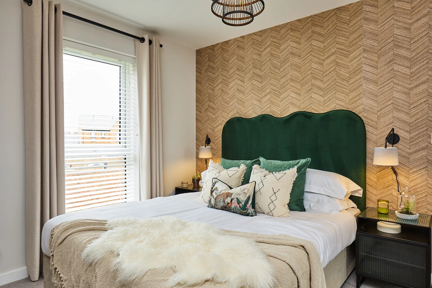A cozy, modern bedroom featuring a green upholstered headboard, layered pillows in neutral and patterned fabrics, and a beige throw with a white fur accent. The wall behind the bed is covered in textured chevron wallpaper. Two bedside tables hold lamps and decorative items. A large window with white blinds and beige curtains lets in natural light. A contemporary ceiling light fixture completes the warm, inviting space.