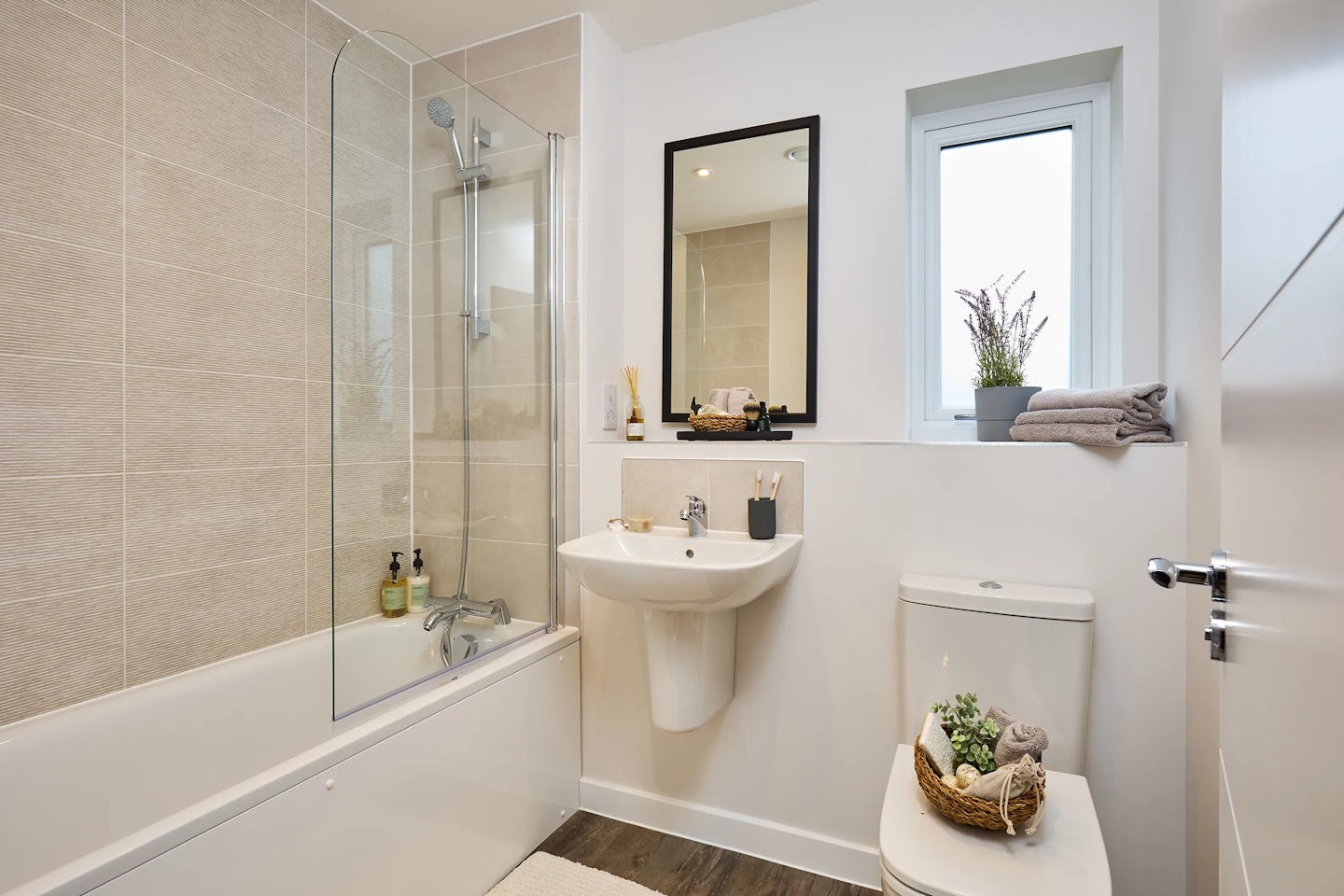 A modern bathroom with light tiled walls and wooden flooring. It features a bathtub with a glass shower screen and wall-mounted showerhead, a white sink with a mirror above, and a toilet topped with a wicker basket holding a plant and decor. A small window above the toilet displays folded towels and a potted plant, adding a touch of greenery. The sink area includes a toothbrush holder and soap dispenser, creating a clean, lived-in feel