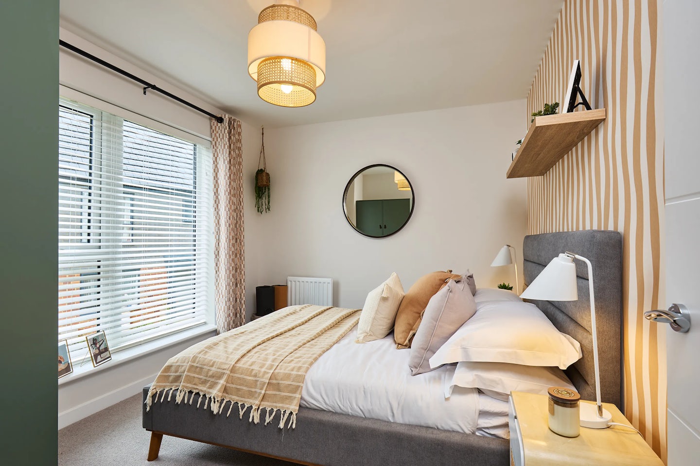 A stylish bedroom with a grey upholstered bed centered against a striped wallpapered wall. The bed is dressed in beige and white bedding with layered pillows and a throw blanket. Above the bed hangs a round mirror and a wooden shelf displaying a small plant and framed picture. A bedside table with a lamp and candle sits to the right. On the left, a large window with white blinds and patterned curtains lets in natural light. Two framed pictures rest on the windowsill, and a hanging plant adds greenery. A woven ceiling light fixture and radiator complete the cozy, modern space
