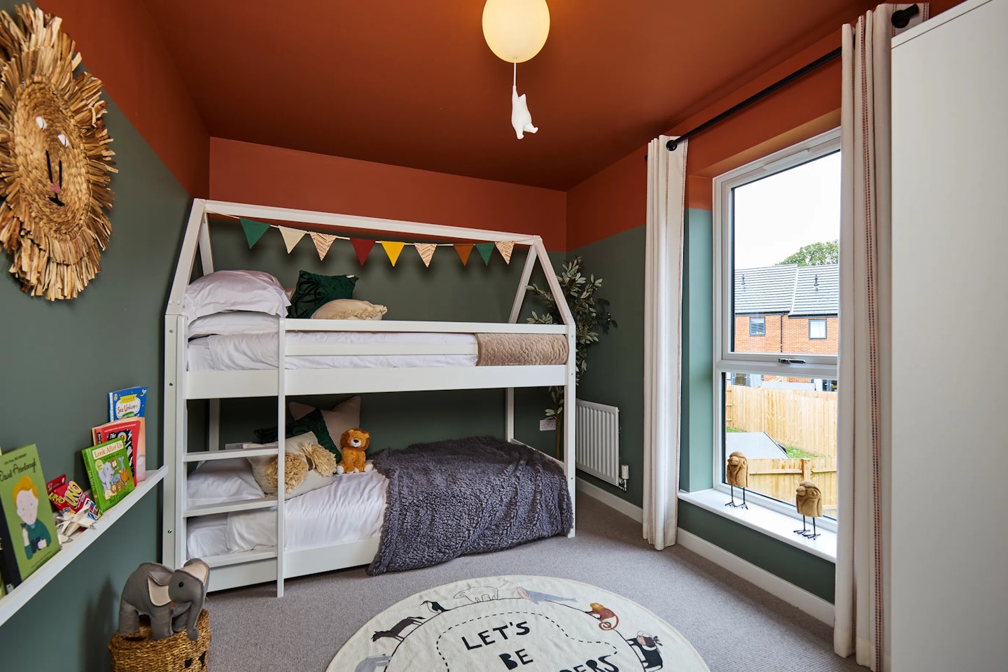 A cozy children’s bedroom with orange and green walls, featuring a white bunk bed. The top bunk is decorated with colorful triangular bunting, while the bottom bunk has a gray blanket and a stuffed lion toy. A round rug with animal illustrations and the words “LET’S BE” sits on the wooden floor. A shelf holds books, toys, and a wicker basket, with an elephant toy on display. Natural light streams through a large window with white curtains above a radiator. A wardrobe stands to the right, and a round ceiling light with a hanging decoration completes the cheerful space.