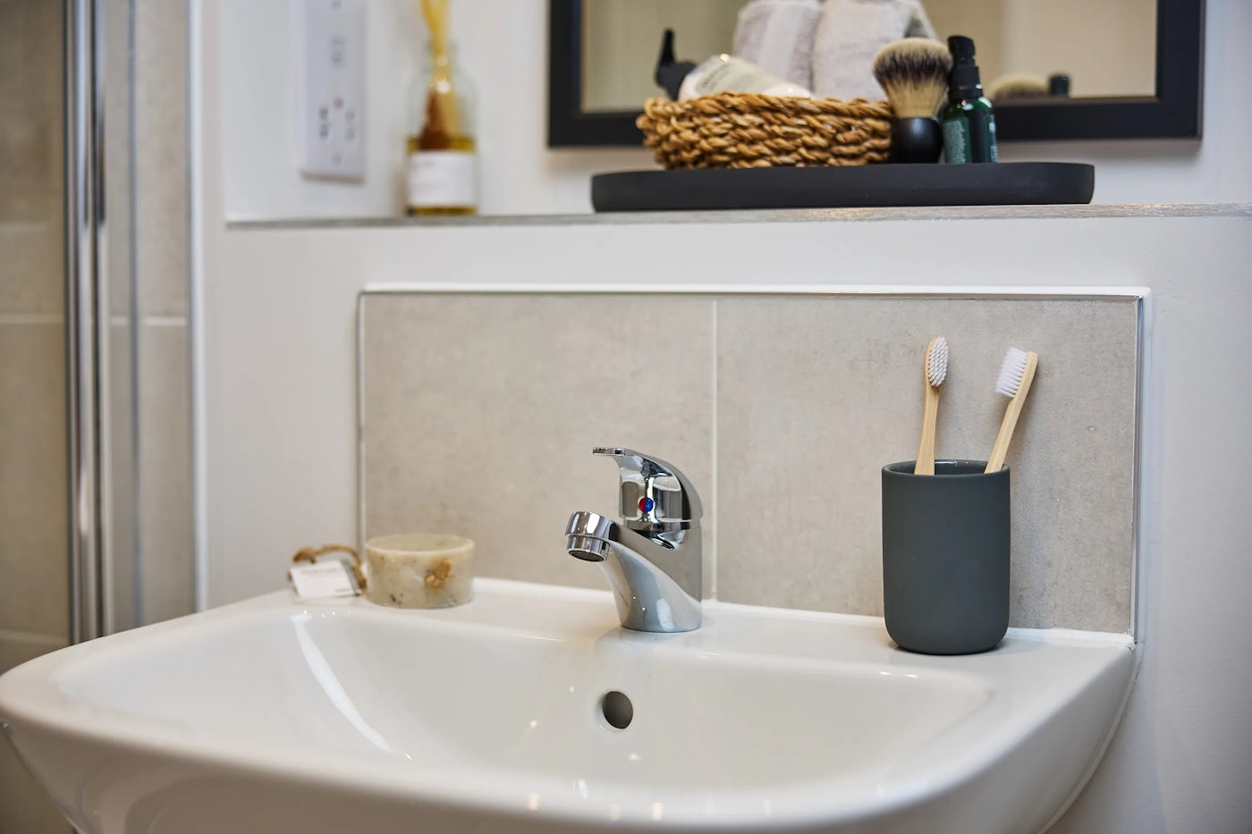 A white bathroom sink with a chrome faucet, flanked by a dark grey toothbrush holder and a soap dish with a bar of soap. Above the sink, a shelf holds a wicker basket filled with toiletries including a shaving brush and bottles. A mirror is mounted on the tiled wall behind the shelf, reflecting the light-toned bathroom decor