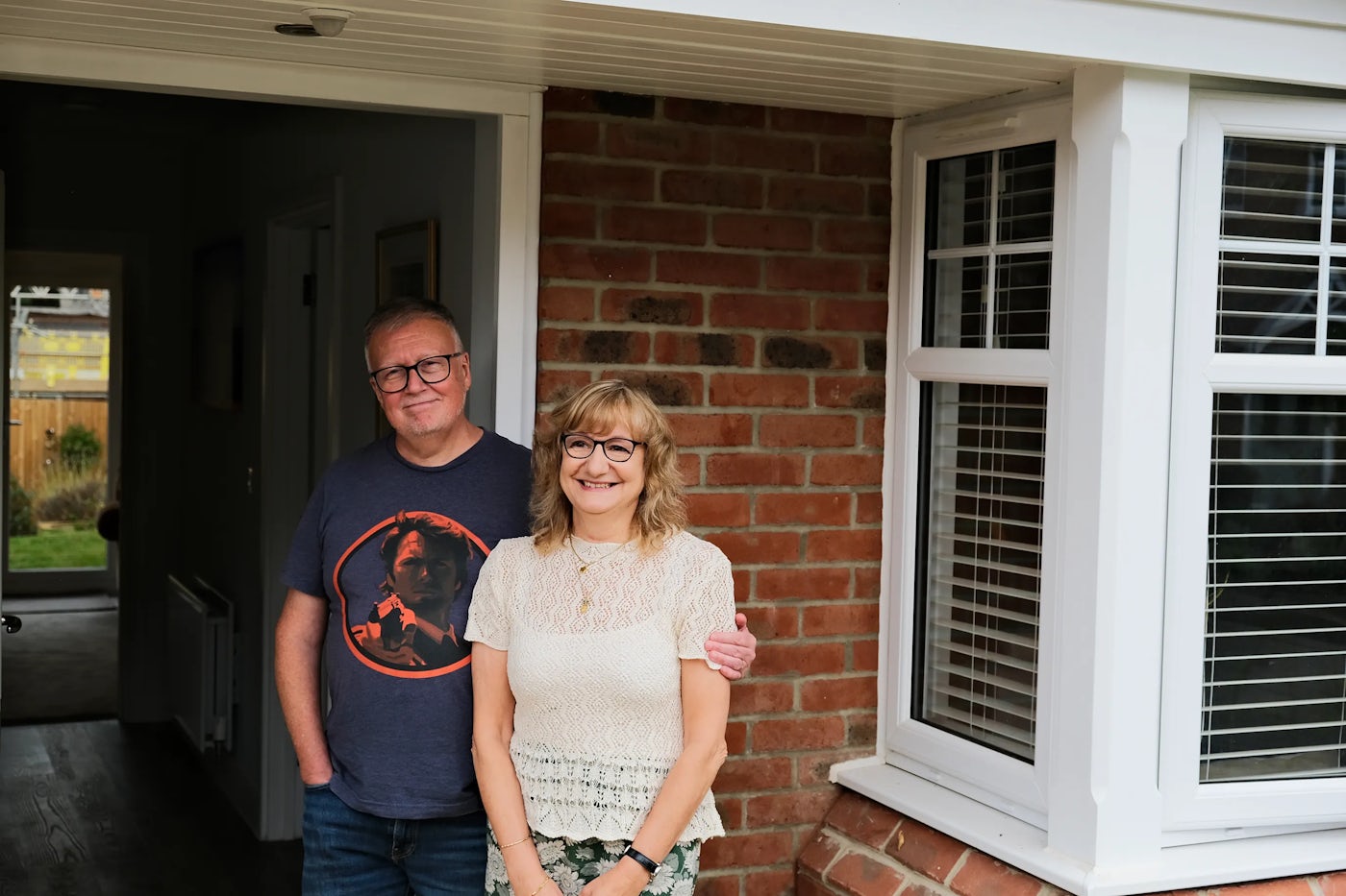 Two people stand outside a brick house with white-framed windows and blinds. The person on the left wears a navy blue t-shirt with an orange graphic and blue jeans, with their arm around the person on the right, who wears a white lace top and patterned trousers. Behind them, an open door reveals a hallway and a glimpse of a garden, suggesting a warm, welcoming moment.