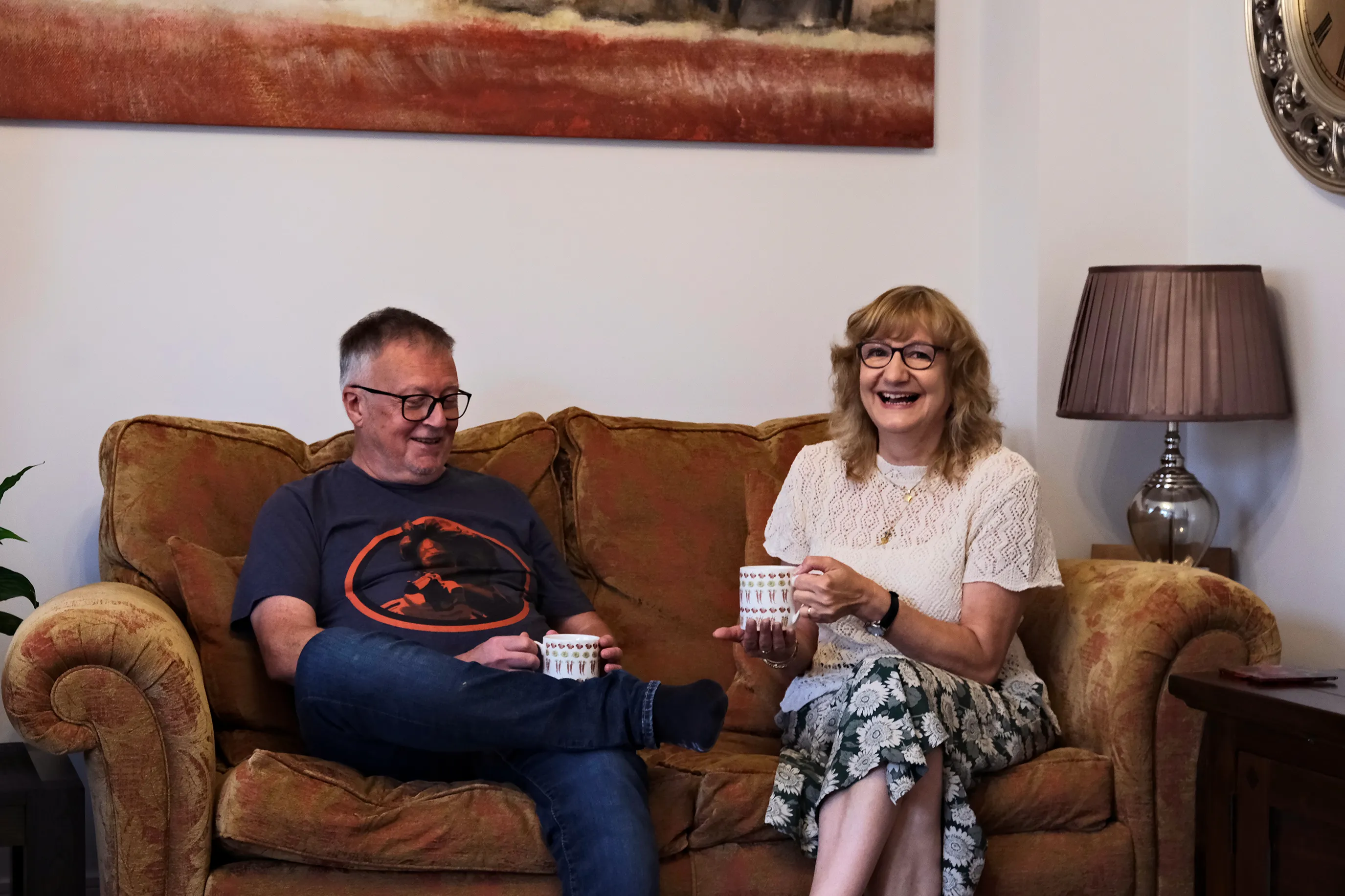 Two people sit on a brown sofa in a cozy living room, each holding a white mug with a red and blue pattern. The person on the left wears a dark t-shirt with an orange graphic and blue jeans; the person on the right wears a white short-sleeved top and a floral skirt. Behind them hangs a large painting in red and brown tones, and a wooden side table with a brown lamp adds to the inviting atmosphere.